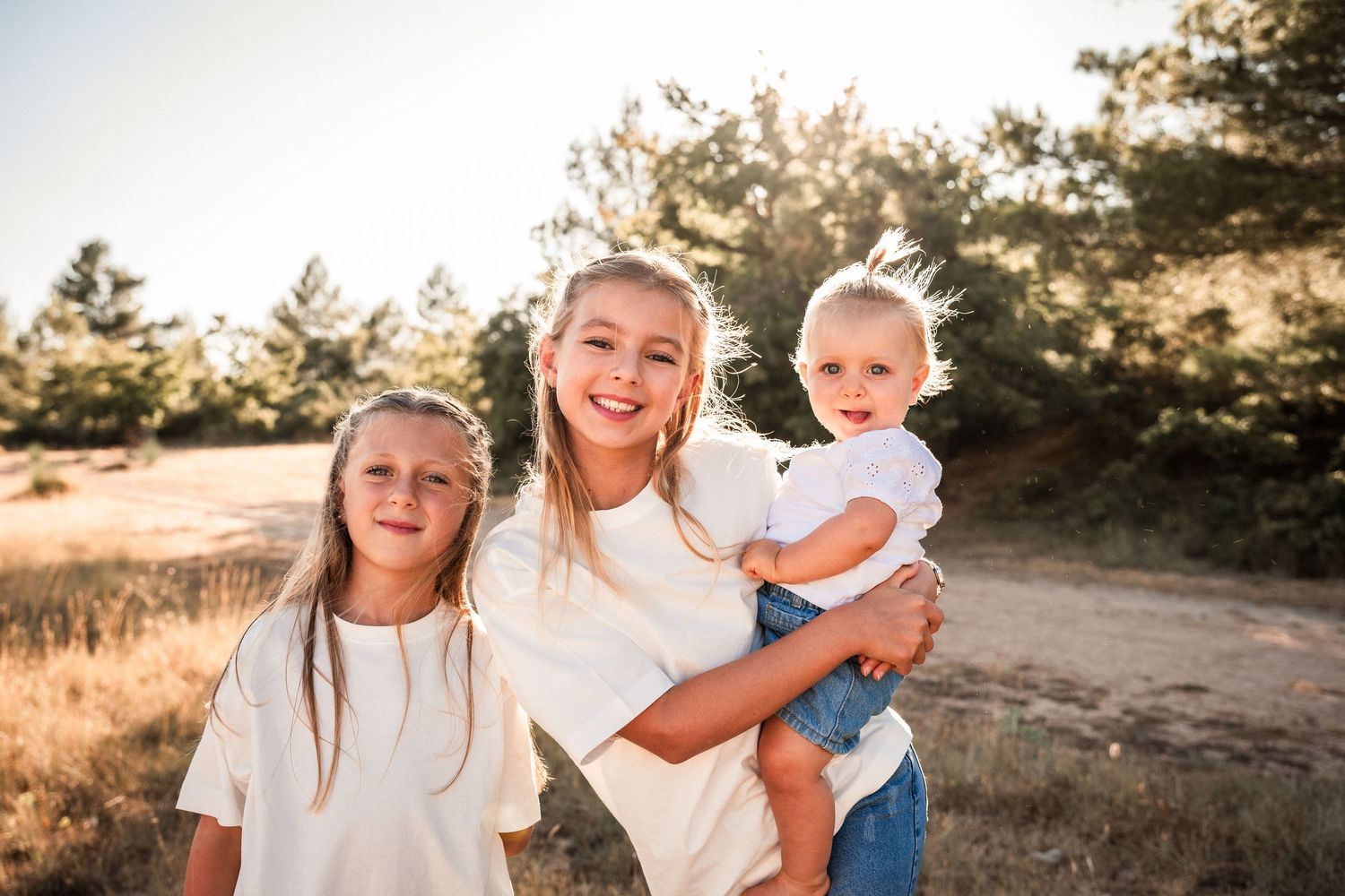 Three young sisters smile outdoors in a sunny field, oldest holding toddler, all wearing white tops.