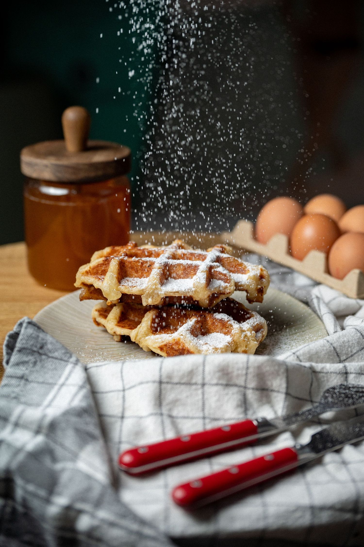 Powdered sugar being dusted over golden Belgian waffles on a plate with eggs and honey.