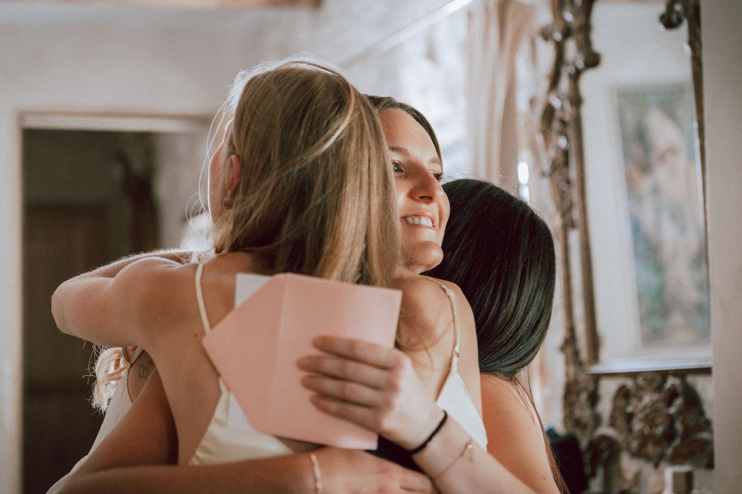 Three women sharing a warm group hug, one holding a pink card, smiling joyfully indoors.