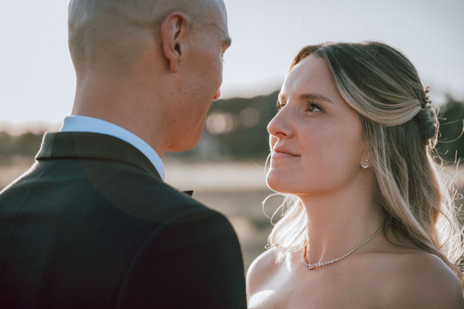Bride gazing lovingly at her groom in close-up portrait with warm golden hour light behind them.