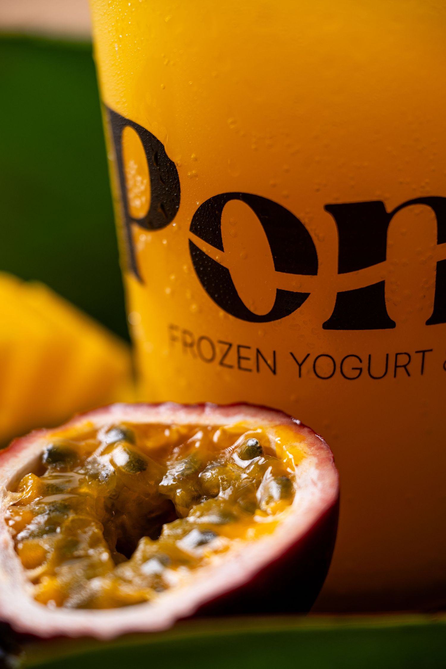 A halved passion fruit in front of a Pom Frozen Yogurt cup, highlighting tropical flavors.