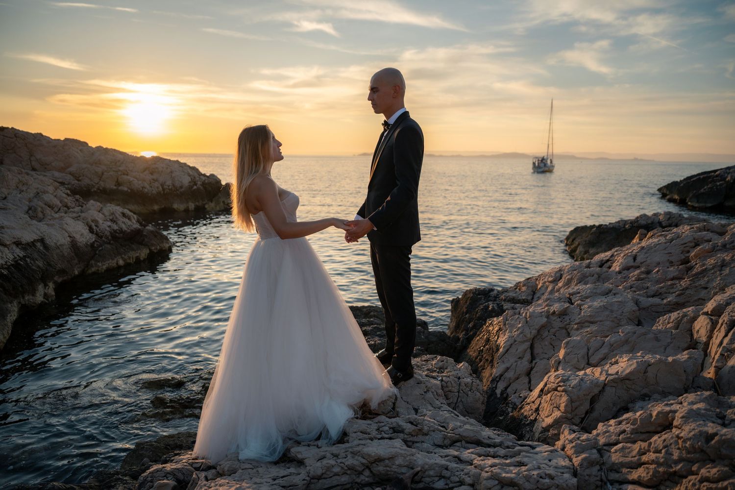 Bride and groom hold hands on rocky shore at sunset with sailboat in background.