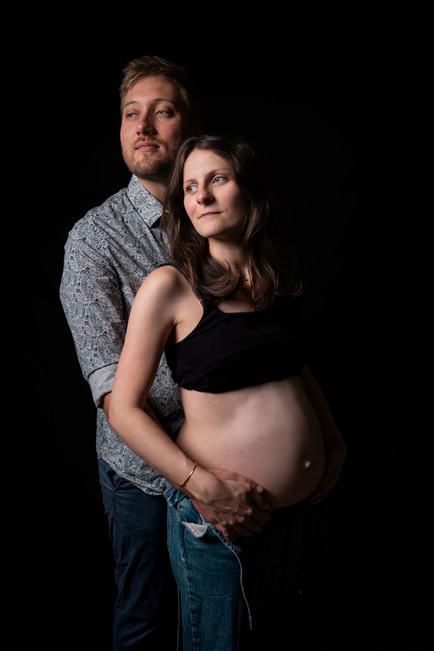 Couple maternity portrait, man embracing pregnant woman from behind, both gazing upward, black background studio photo.