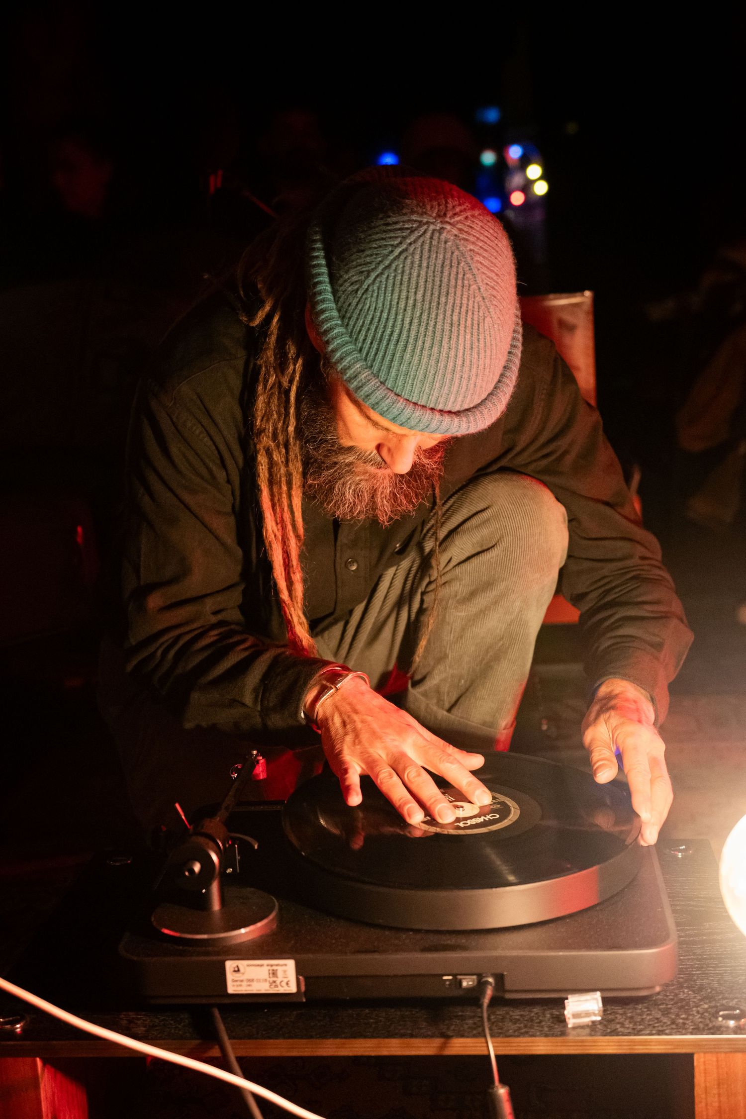 A dreadlocked DJ in a teal beanie spins vinyl records on a turntable under warm stage lighting.
