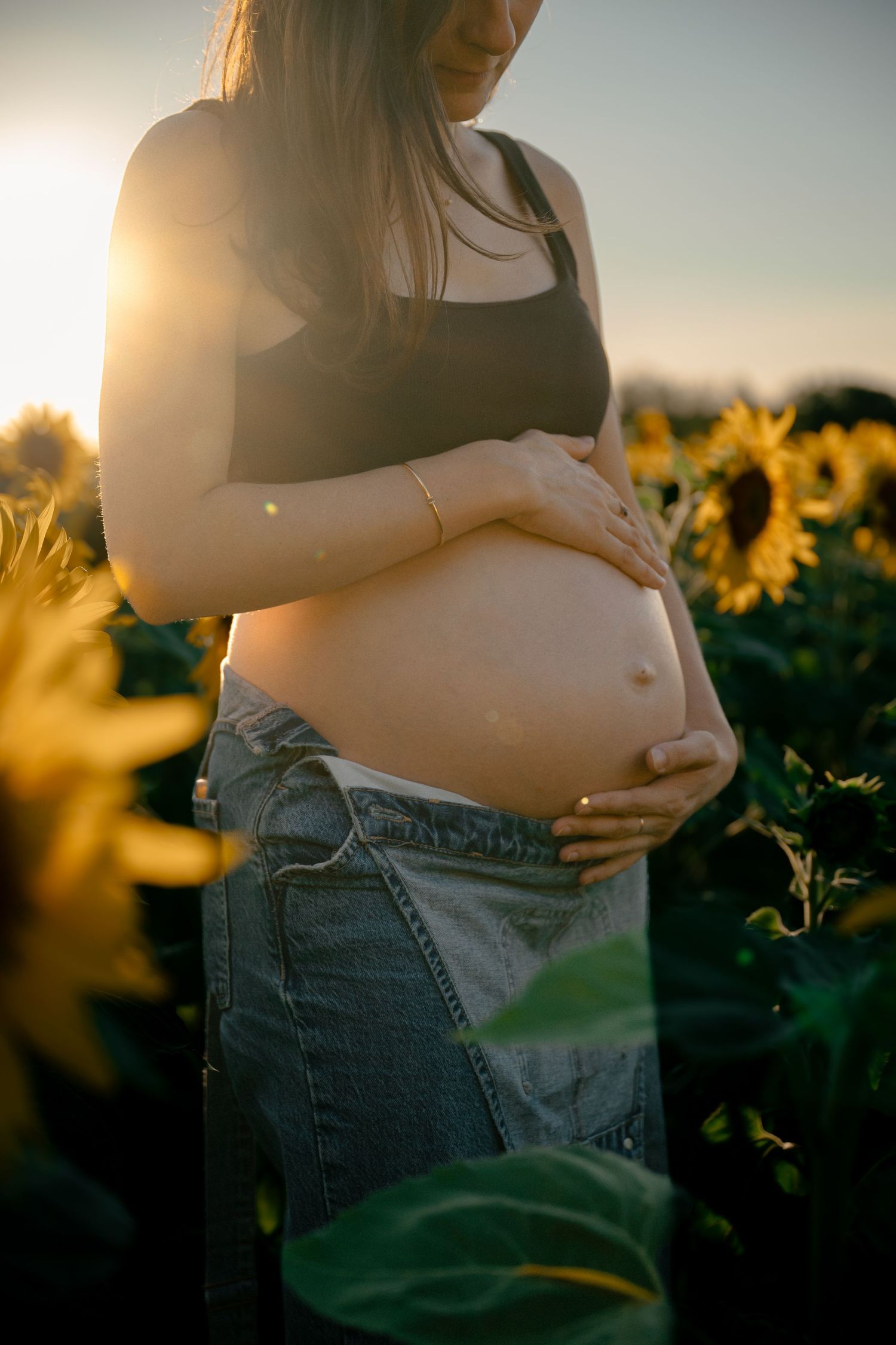 Pregnant woman in black crop top and jeans cradling her baby bump in a sunlit sunflower field at golden hour.