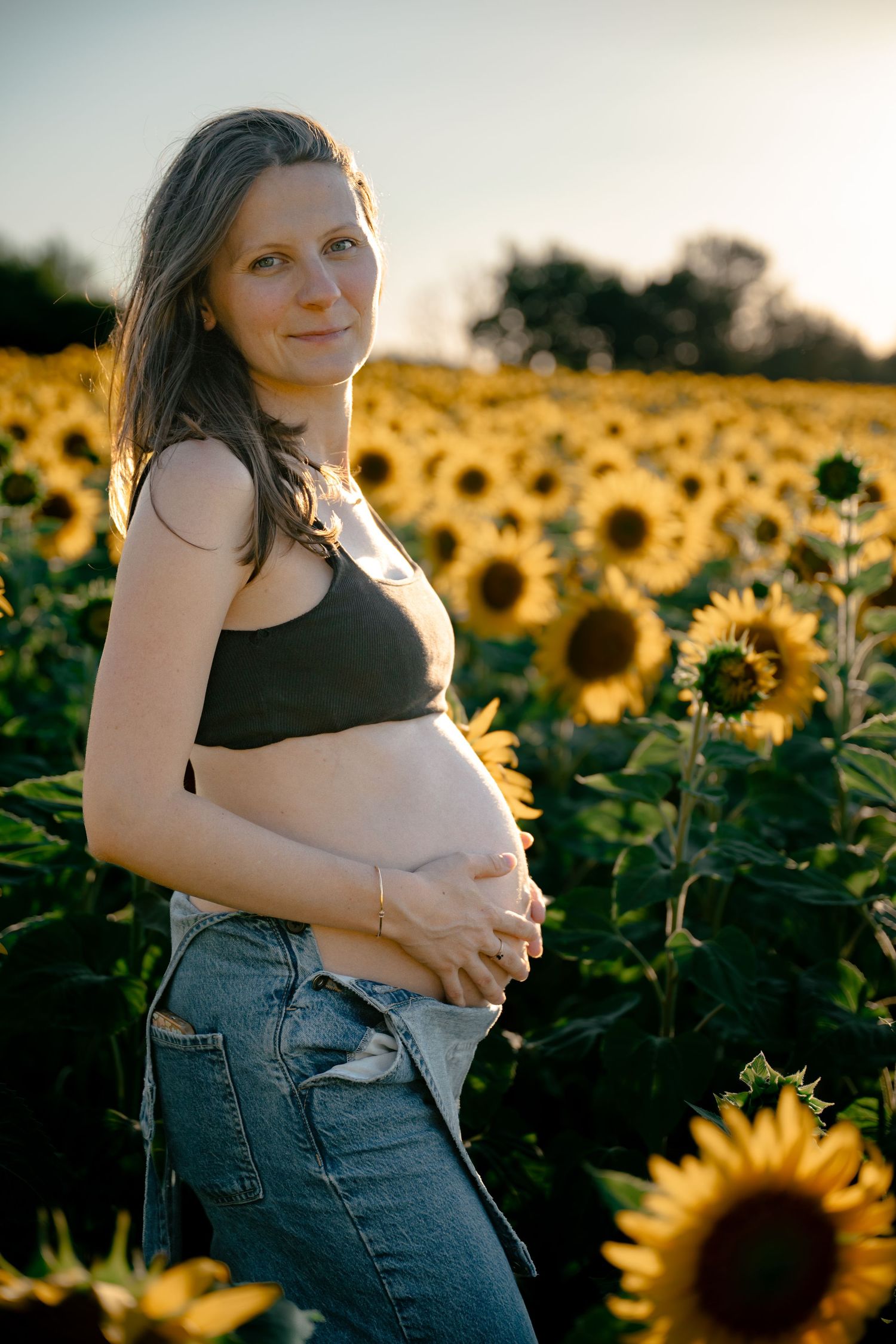 Pregnant woman in black crop top and jeans cradling her baby bump in a sunflower field at golden hour.