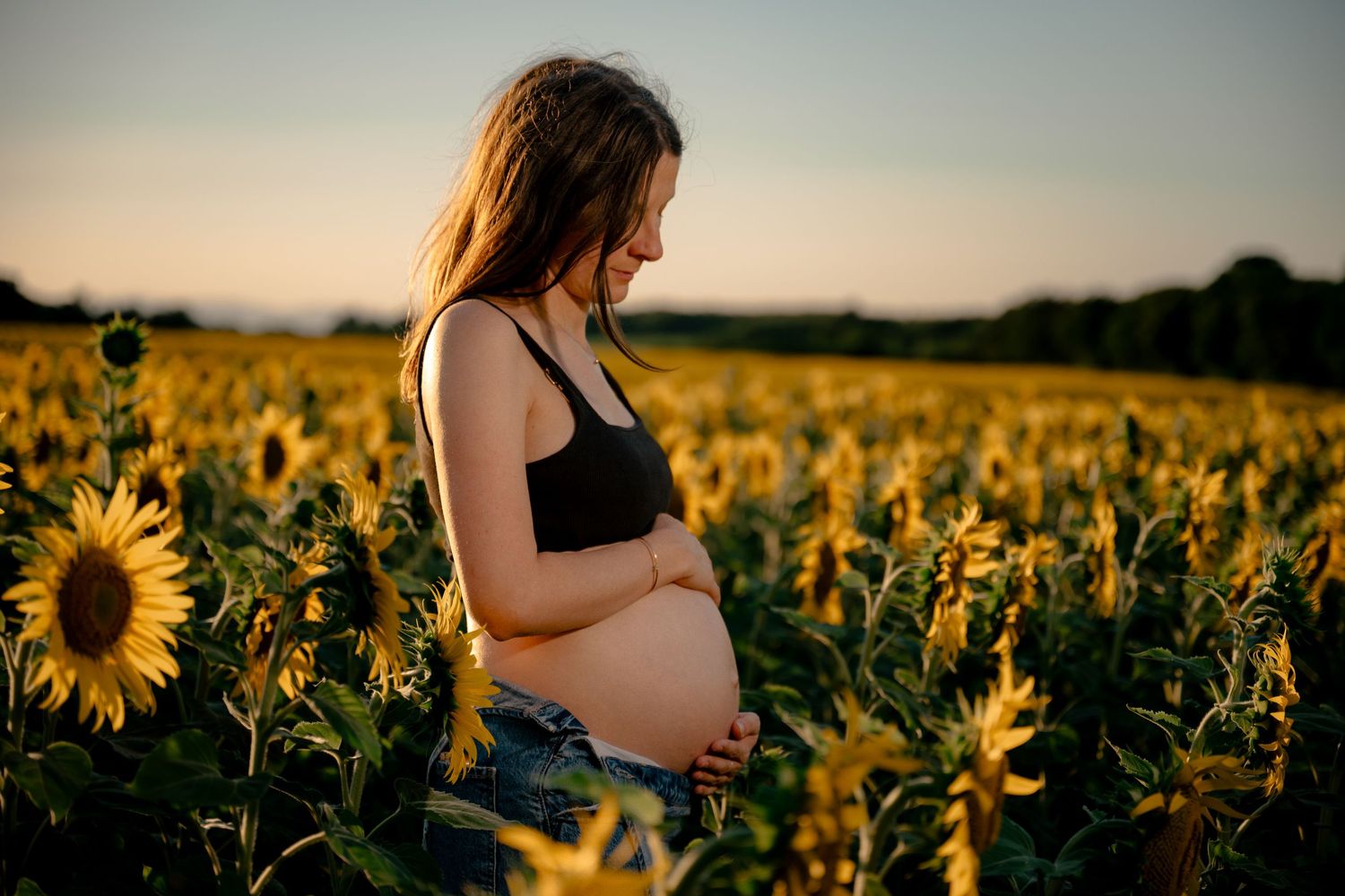 Pregnant woman in black tank top gently holding her baby bump in a sunflower field at golden hour.