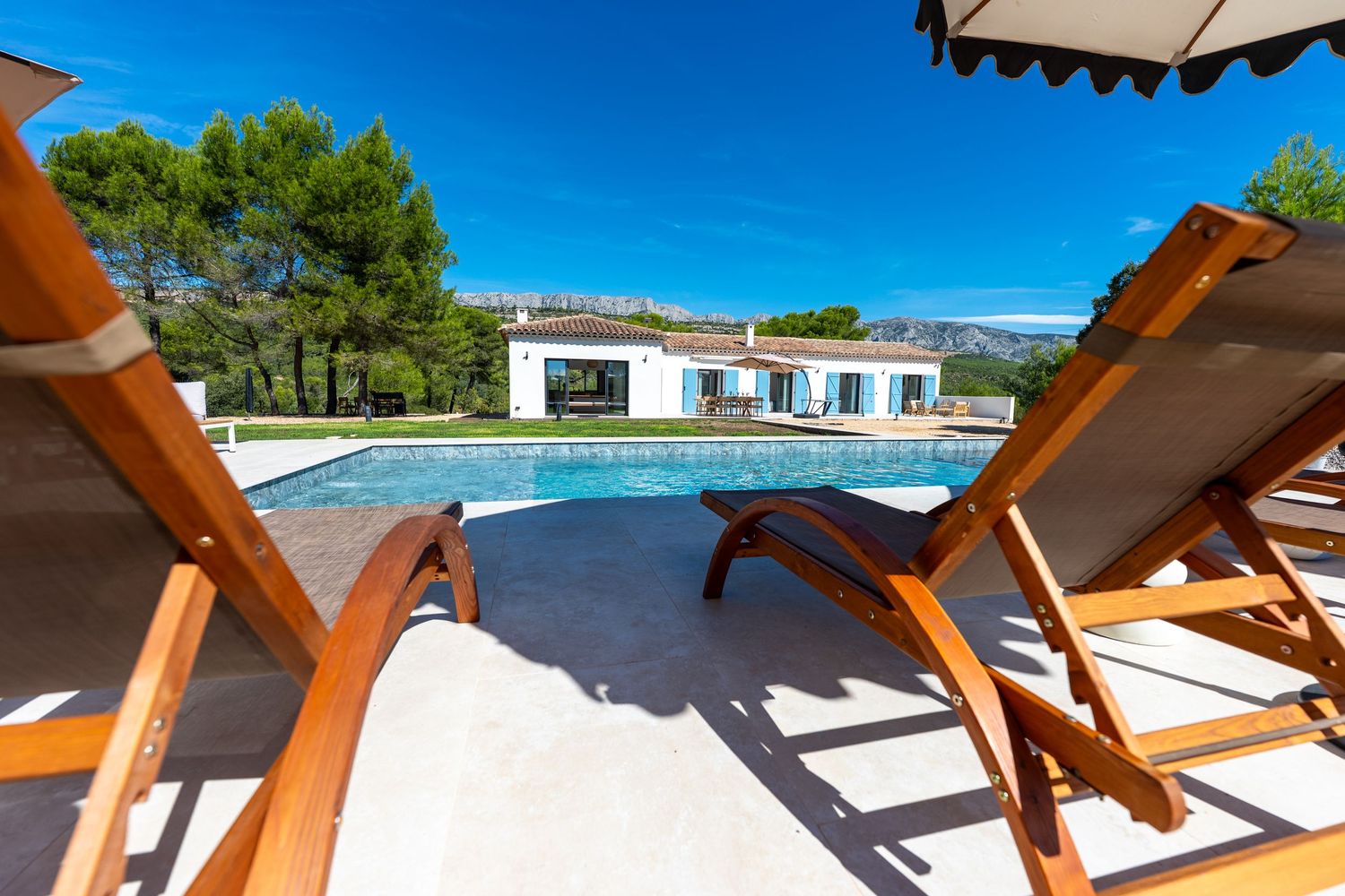 Luxury villa pool with wooden lounge chairs under sunny blue sky surrounded by pine trees.