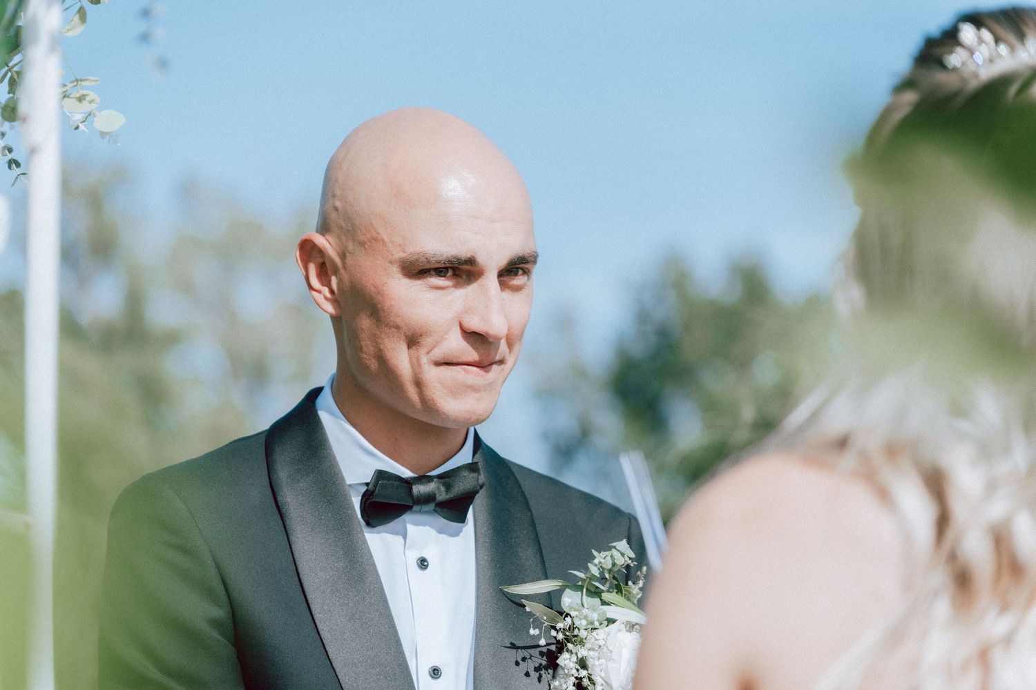 Emotional bald groom in black bow tie tuxedo listens to bride during wedding ceremony.