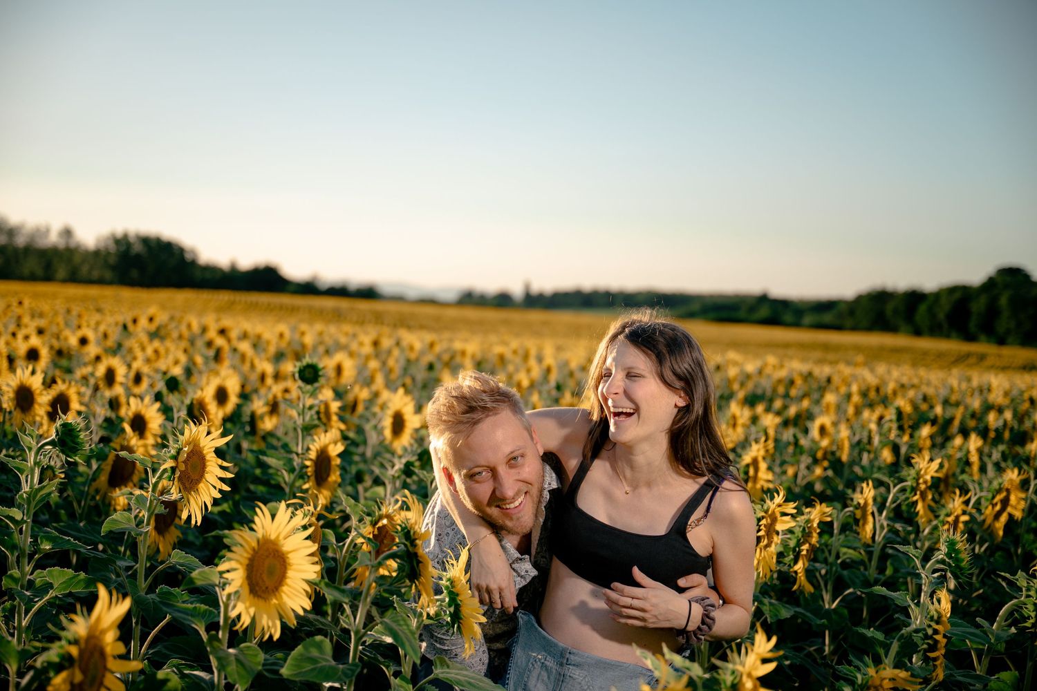 Happy couple laughing together in a vibrant sunflower field during golden hour outdoor maternity shoot.