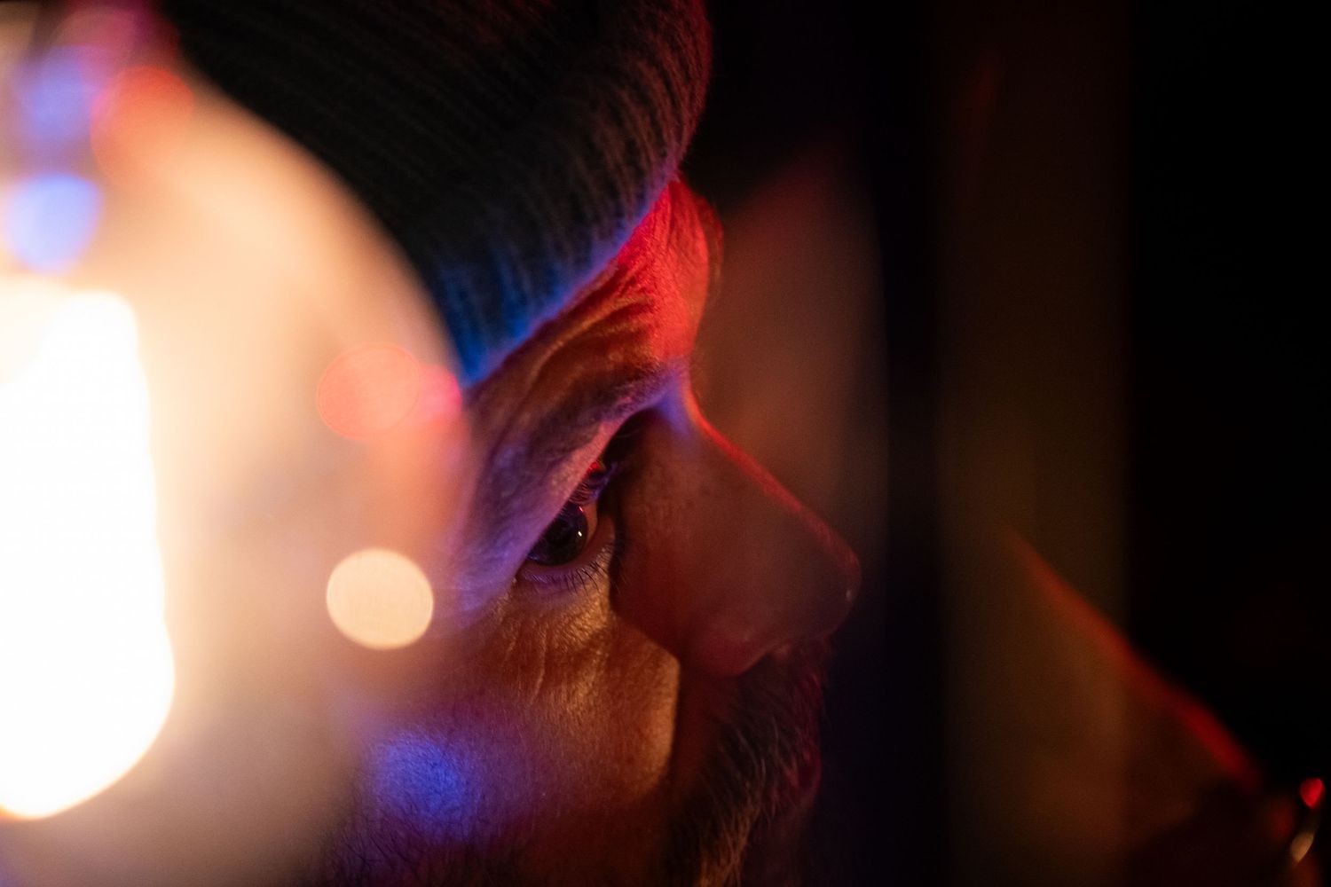 Elderly person in dark beanie hat illuminated by colorful bokeh lights in dramatic close-up portrait.