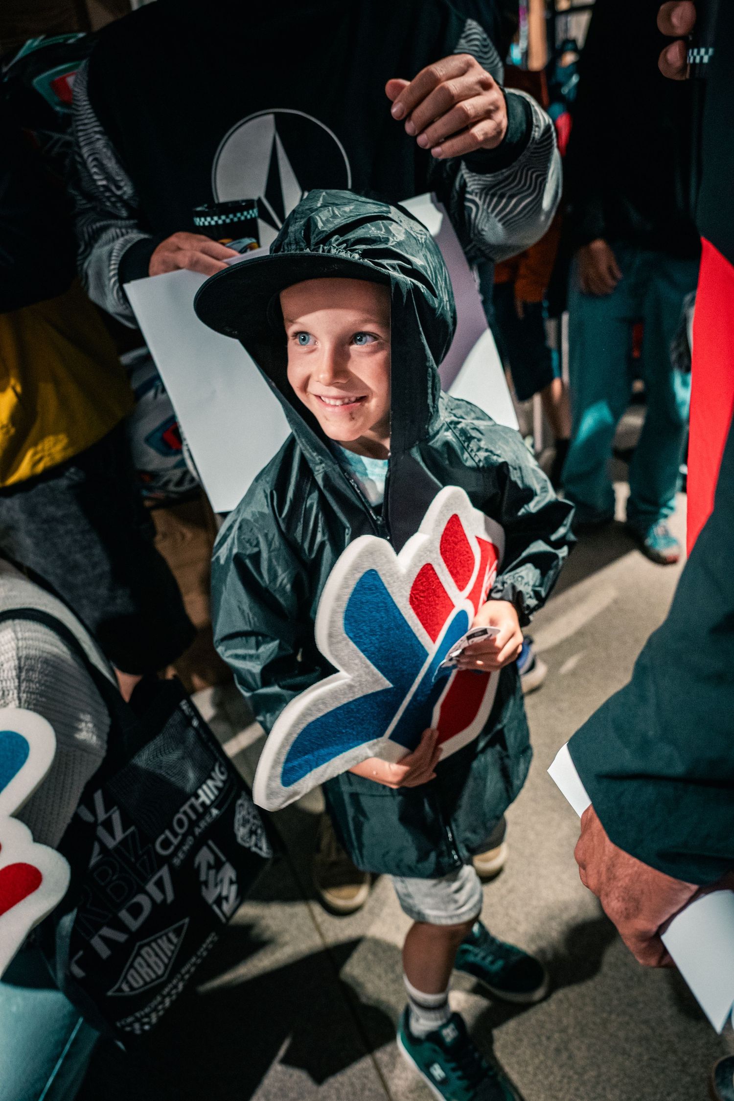 A smiling child in a green raincoat and hat holds a colorful logo cutout at an outdoor sporting event.