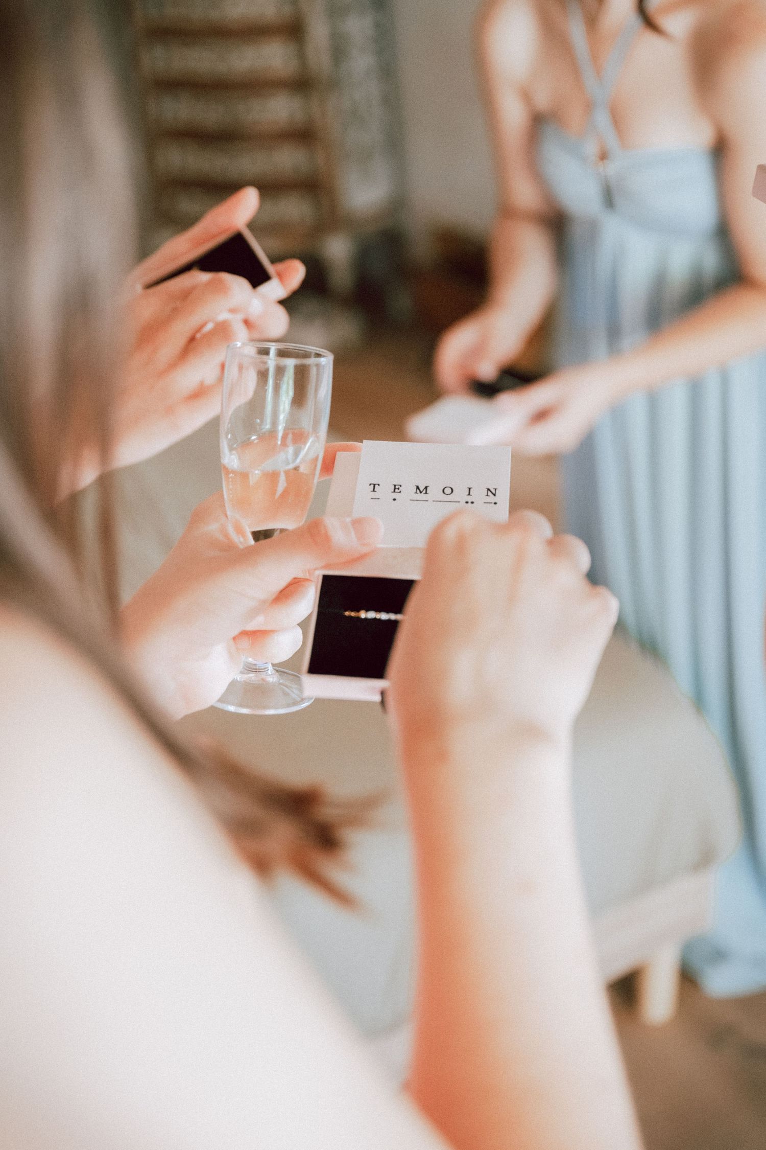 Bridesmaid holding champagne glass and TEMOIN card with jewelry box at wedding preparation.