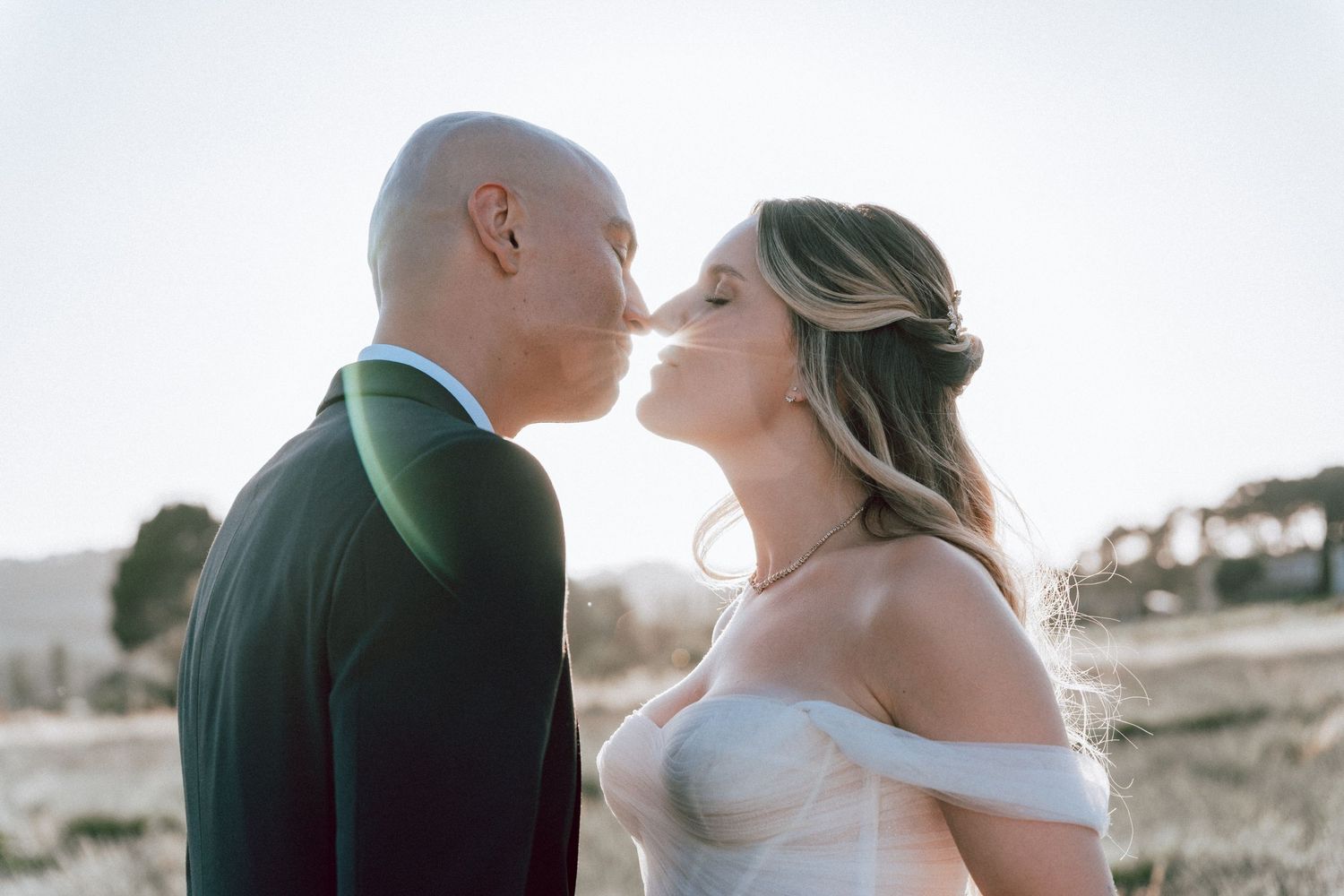 Bride and groom sharing a romantic almost-kiss outdoors in golden sunlight on their wedding day.