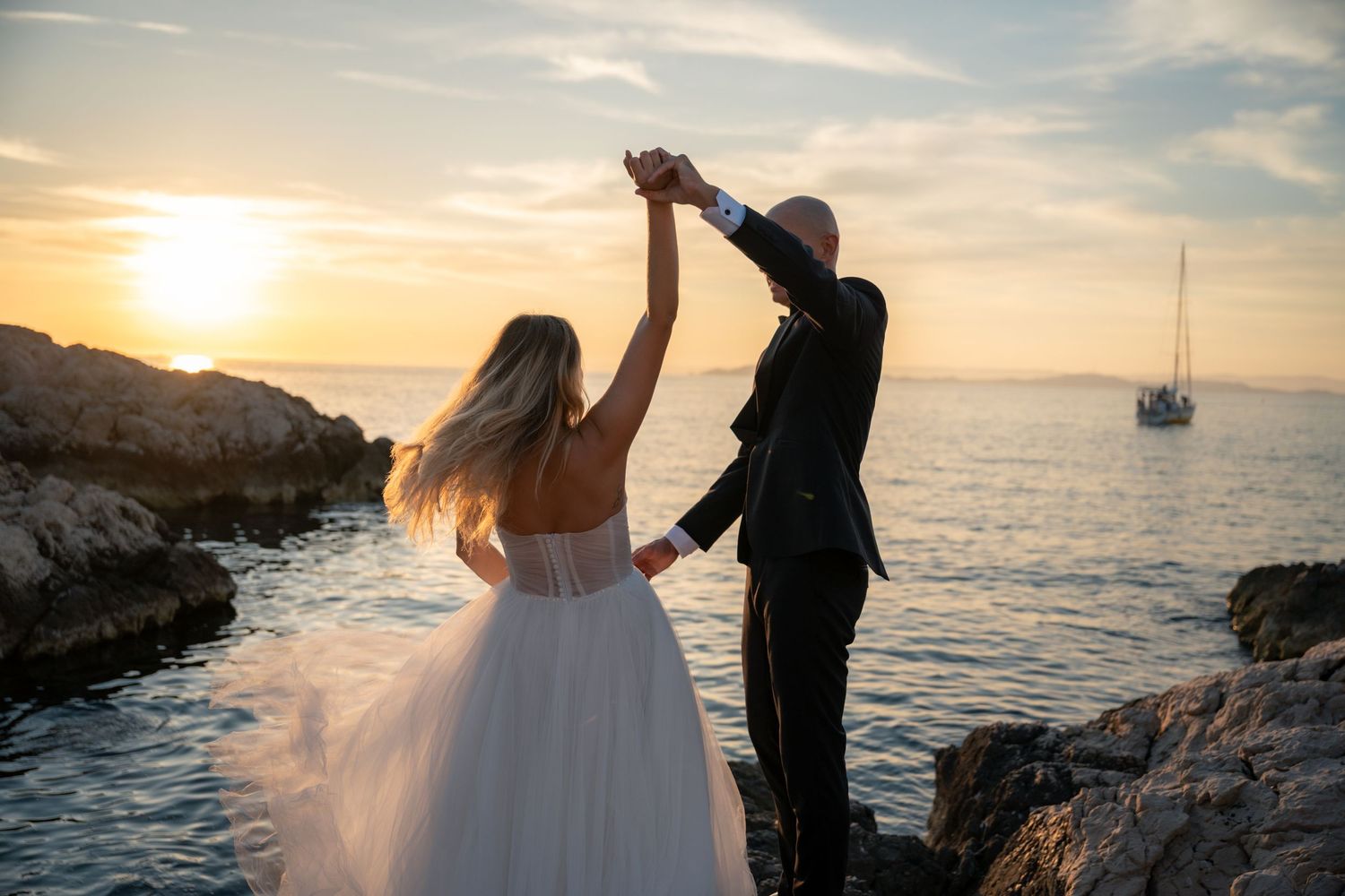 Groom spinning bride in white gown on rocky coast at vibrant sunset.
