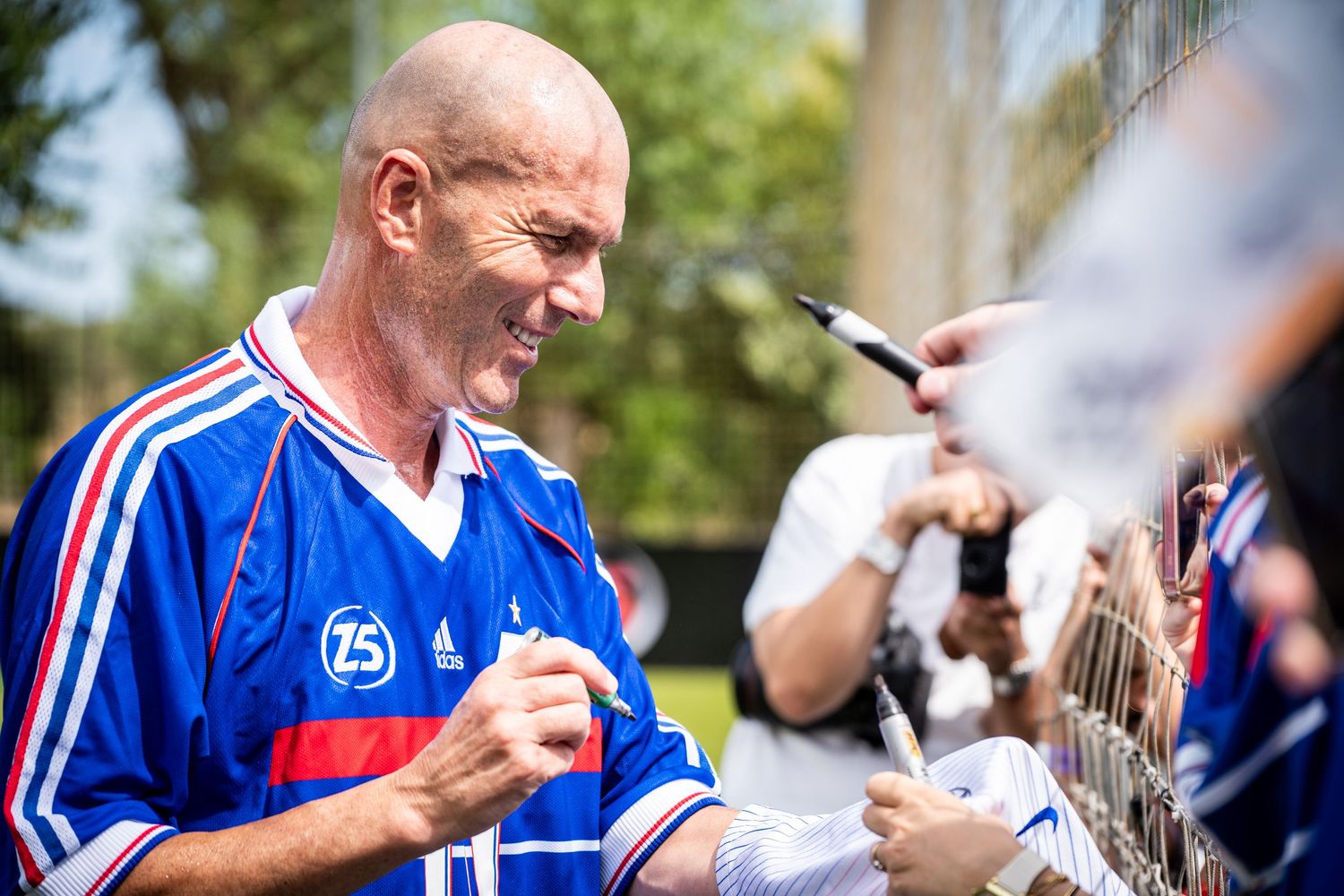 Man in blue French football jersey signing autographs for fans outdoors on a sunny day.