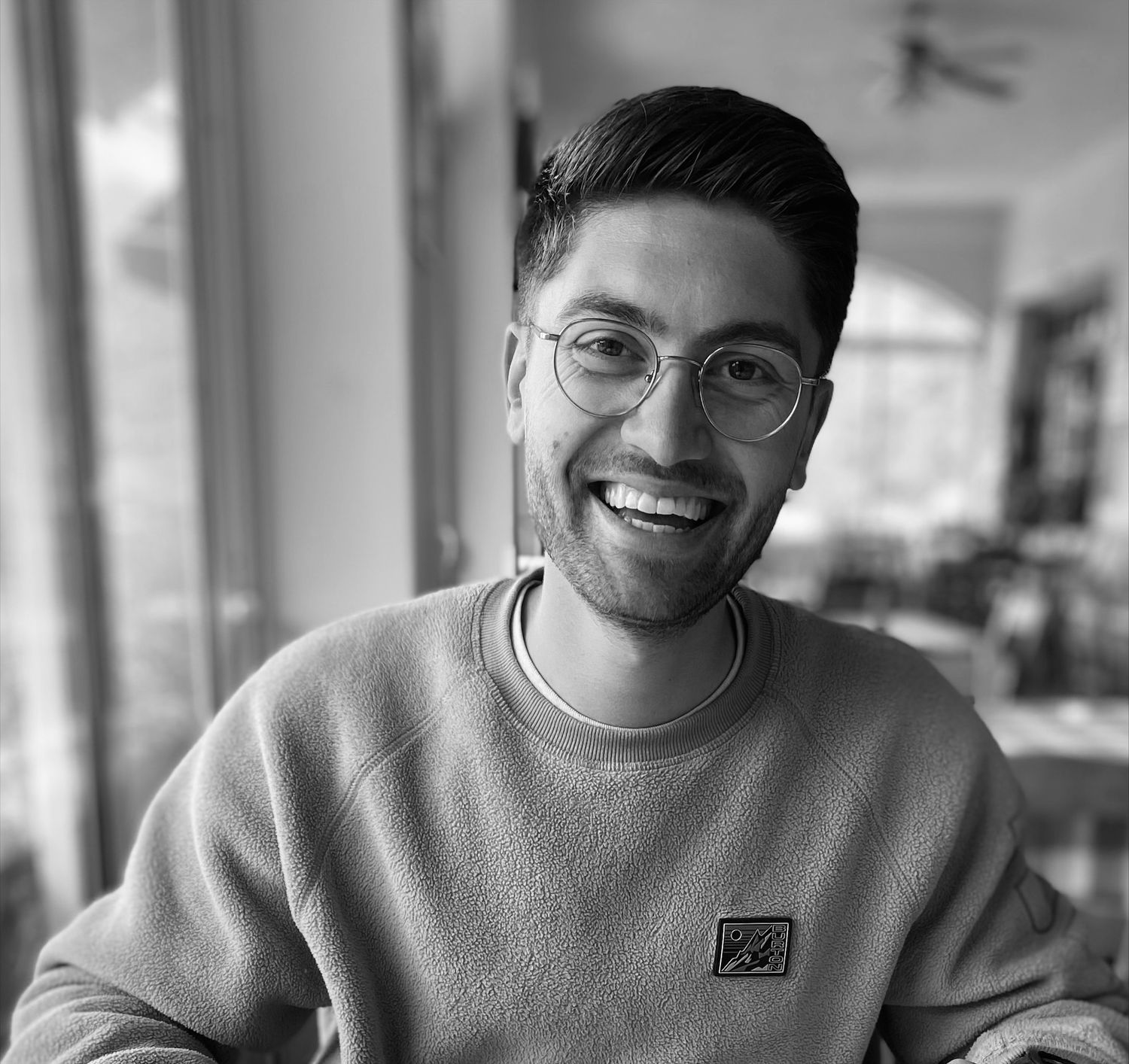 Smiling young man with glasses wearing a casual sweatshirt in a bright indoor setting, black and white photo.