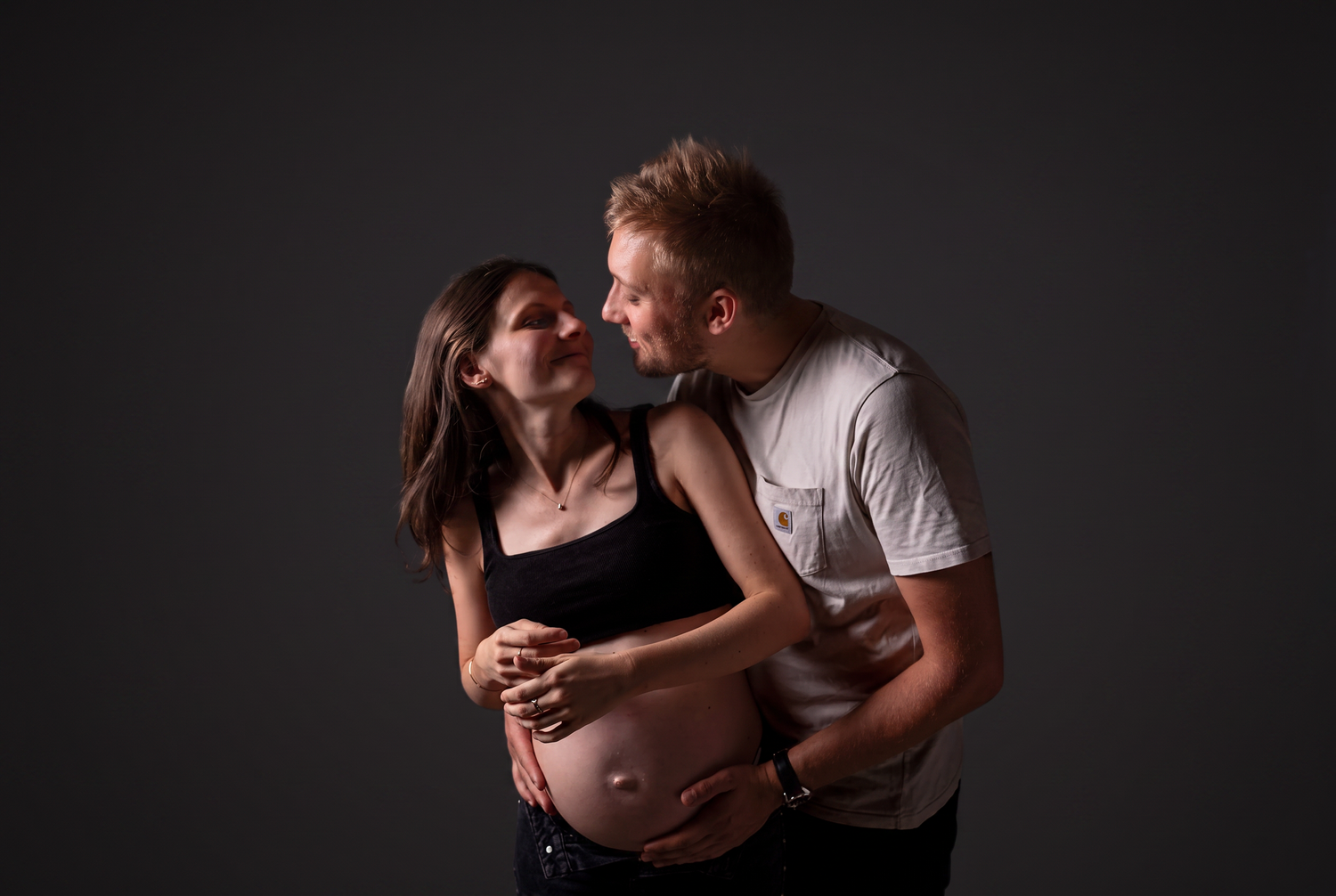 Couple sharing a tender moment, pregnant woman cradling baby bump with partner embracing her in a studio portrait.
