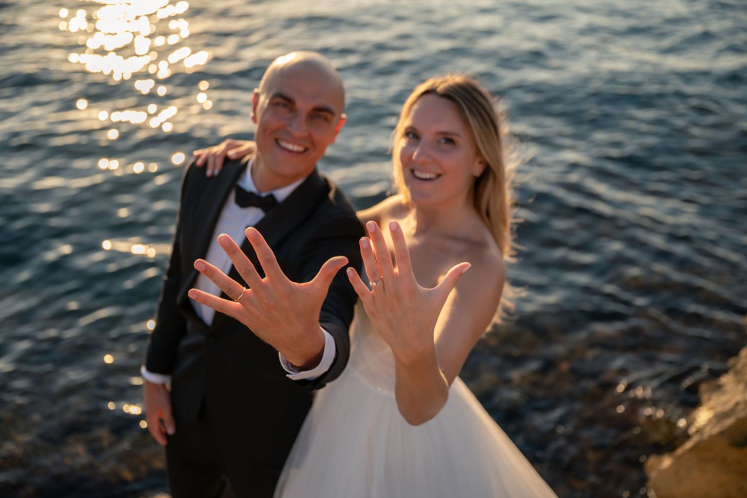 Newlyweds showing wedding rings to camera with sparkling ocean in background.