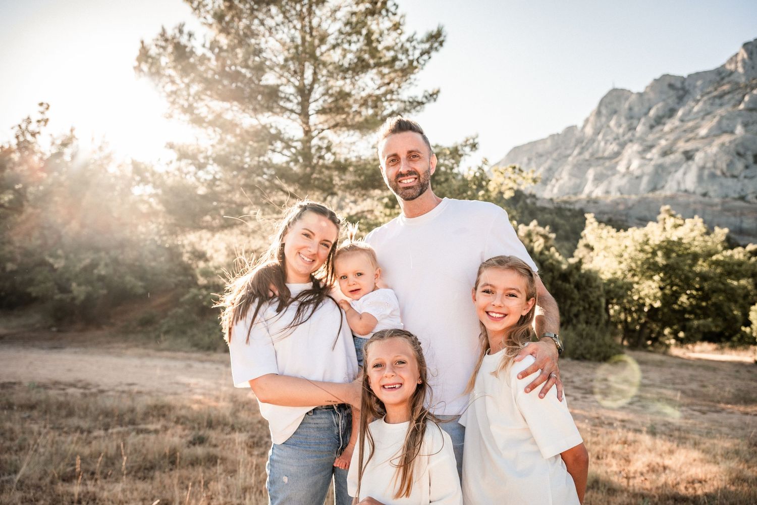 Happy family of five posing outdoors in golden sunlight with mountain and trees in background.