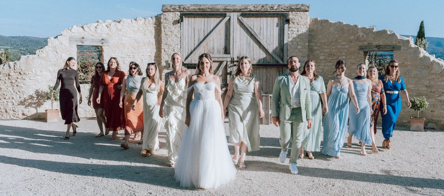 Bride in white gown leading wedding party guests walking together at rustic outdoor venue.