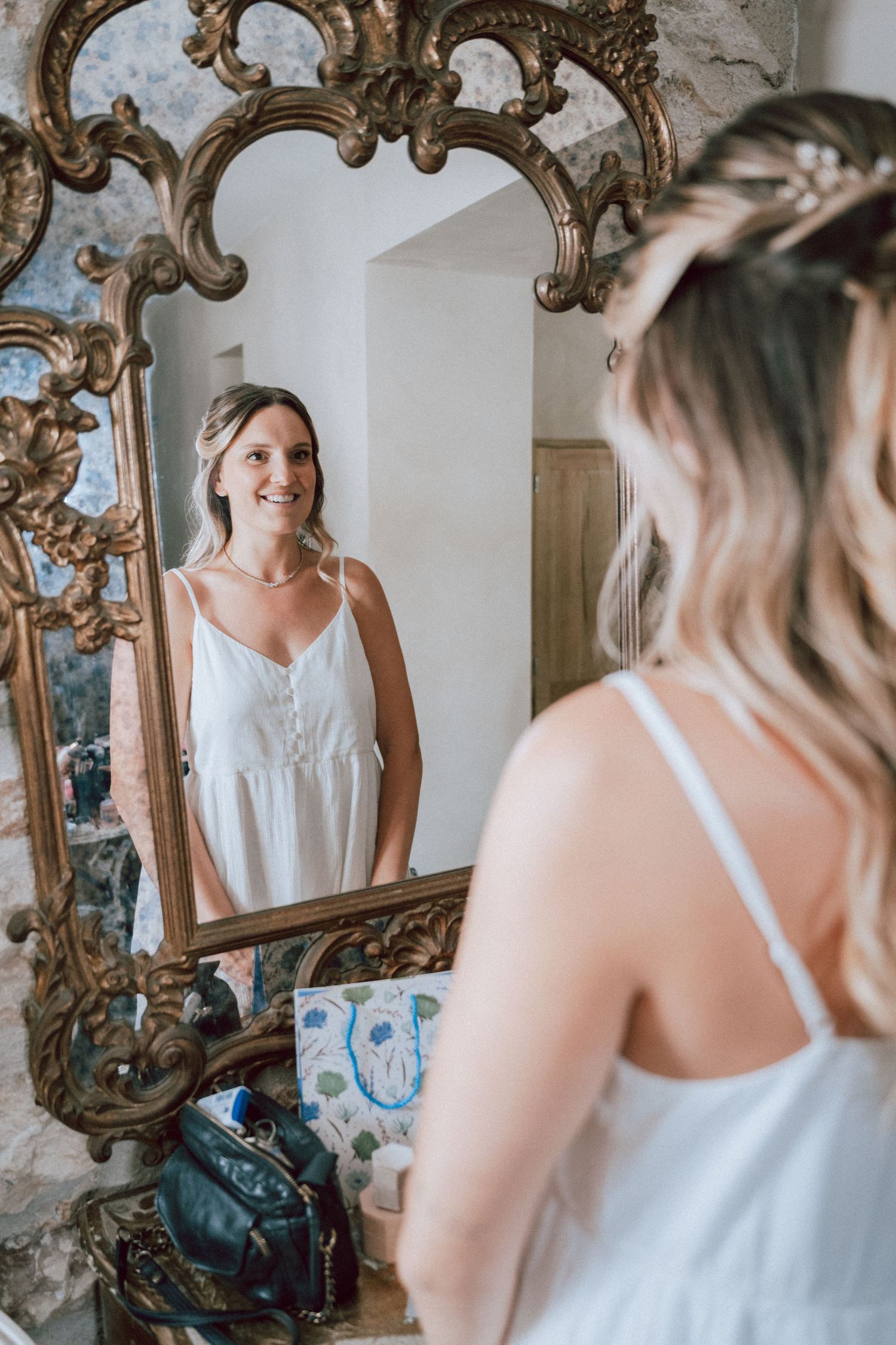 Bride in white dress smiling at her reflection in an ornate gold mirror while getting ready.