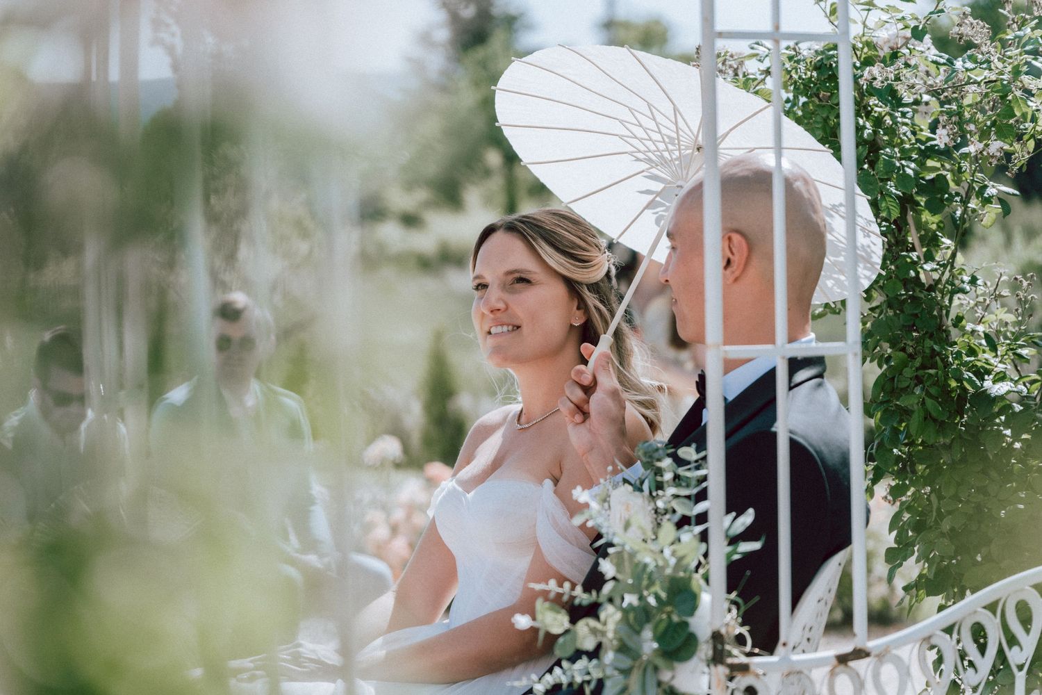 Smiling bride holding white parasol standing beside groom at elegant outdoor wedding ceremony.