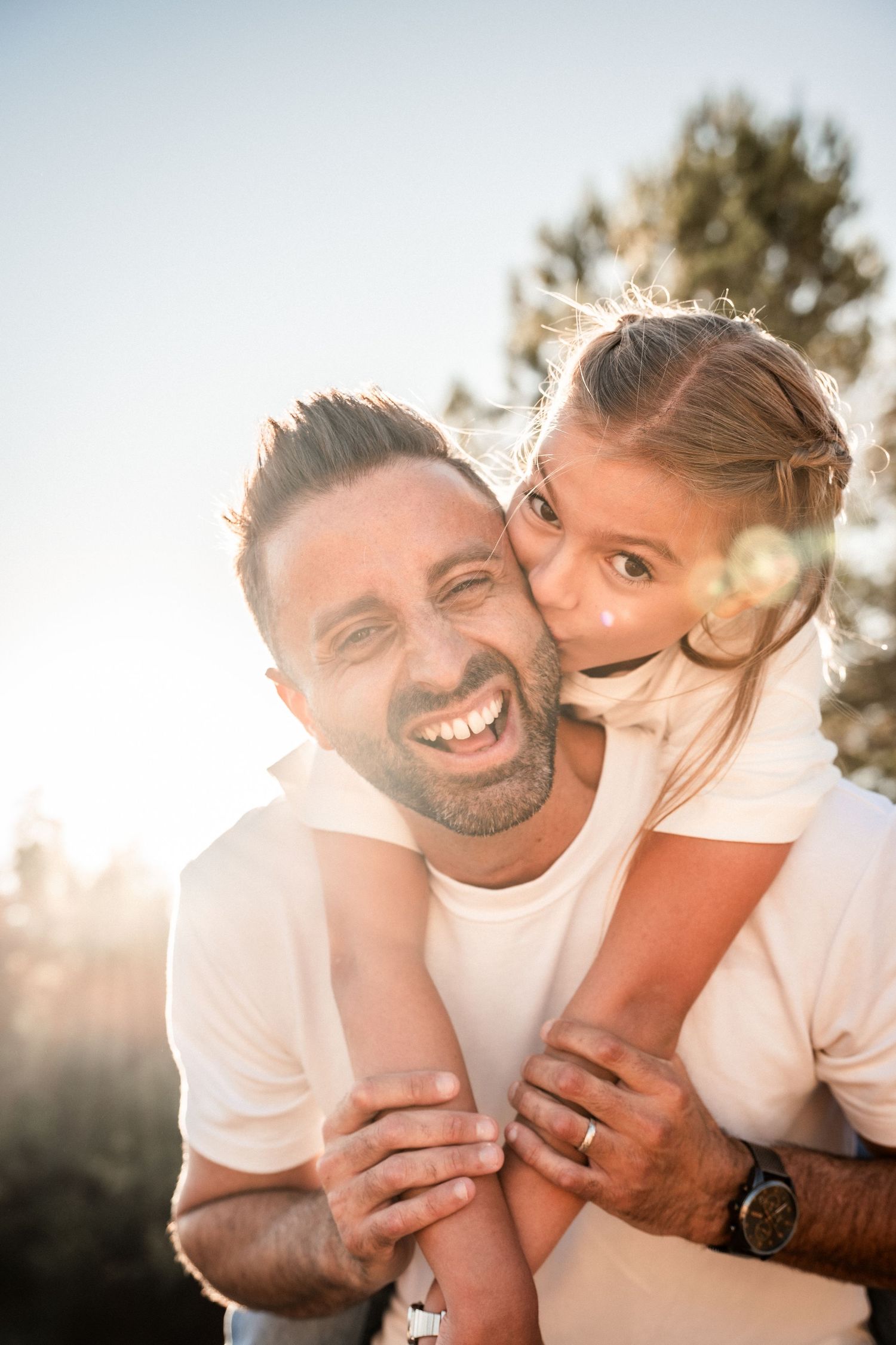 Laughing father receiving a kiss on the cheek from his young daughter outdoors at sunset.