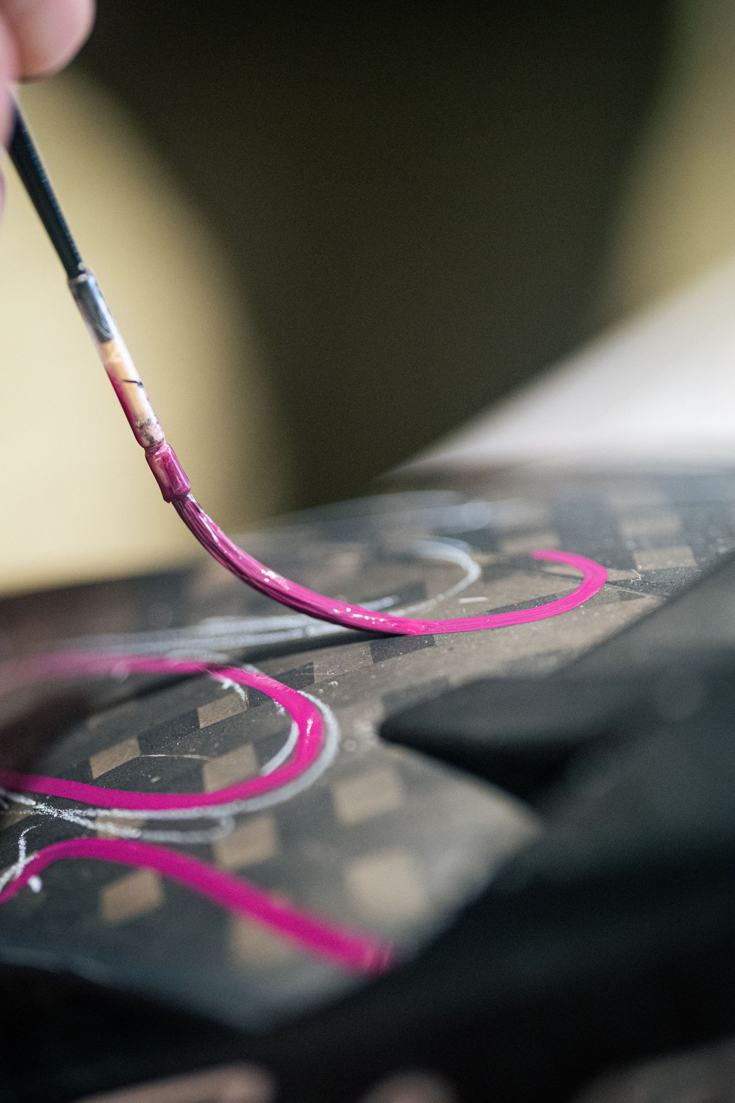 Close-up of a thin paintbrush applying bright pink paint in circular strokes on a dark decorated surface.