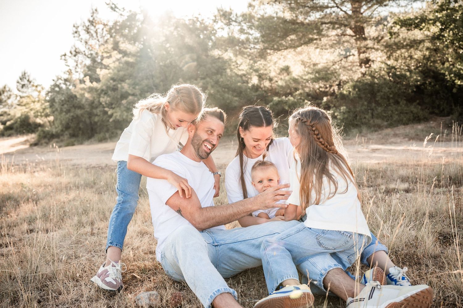 Happy family of five sitting together outdoors in a sunny field, sharing a joyful moment with their young child.