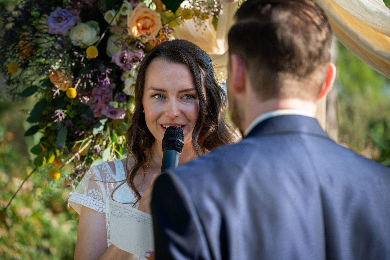 Bride reciting vows into microphone at outdoor wedding ceremony with floral arch.