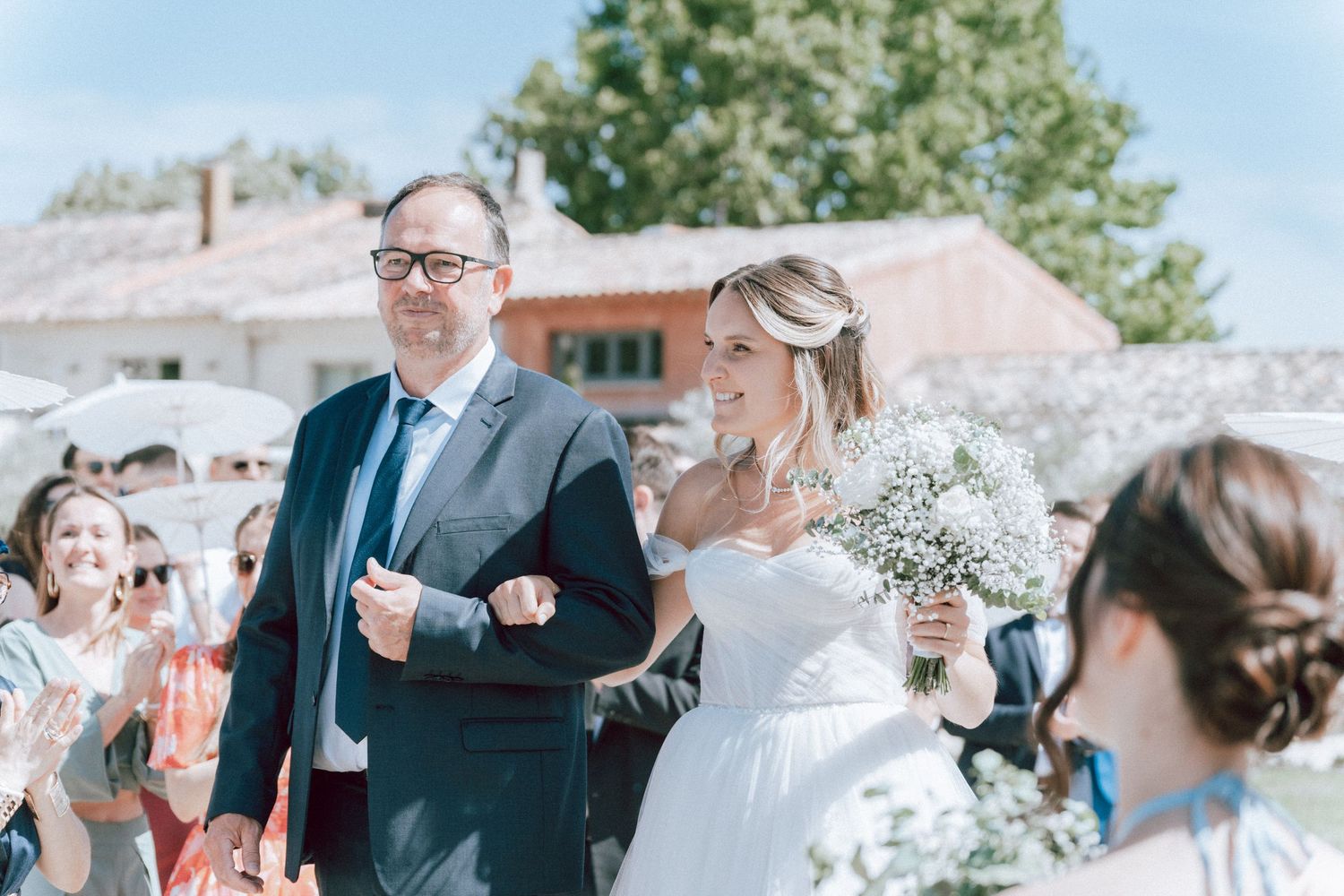 Bride holding white bouquet walks with escort at sunny outdoor wedding ceremony while guests applaud.