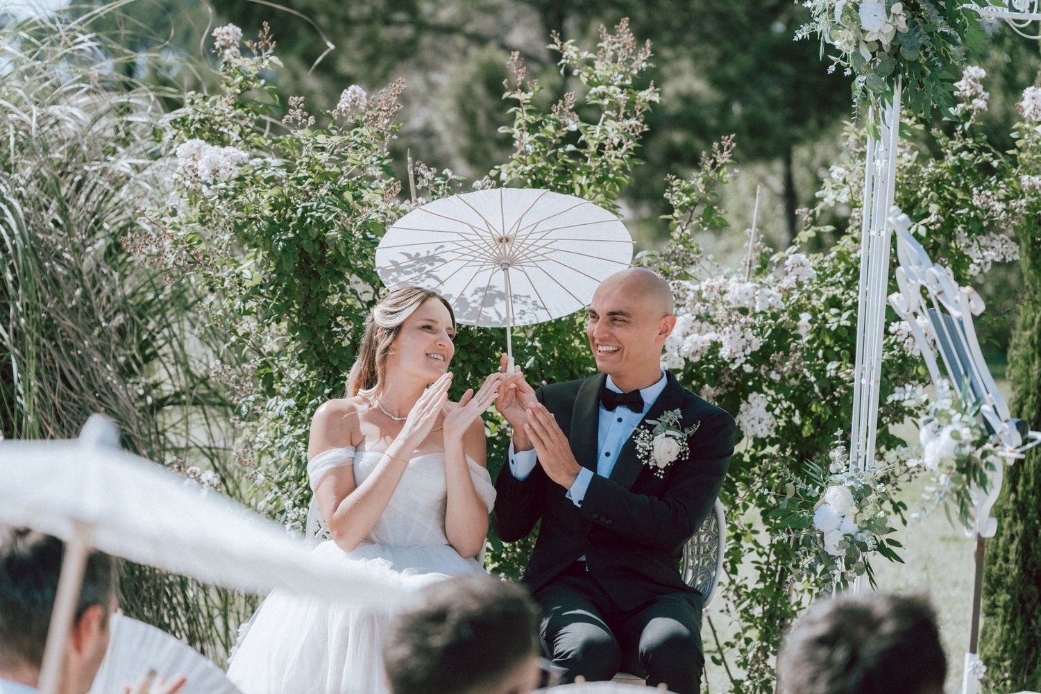 Bride and groom share parasols and smile during outdoor wedding ceremony surrounded by greenery.