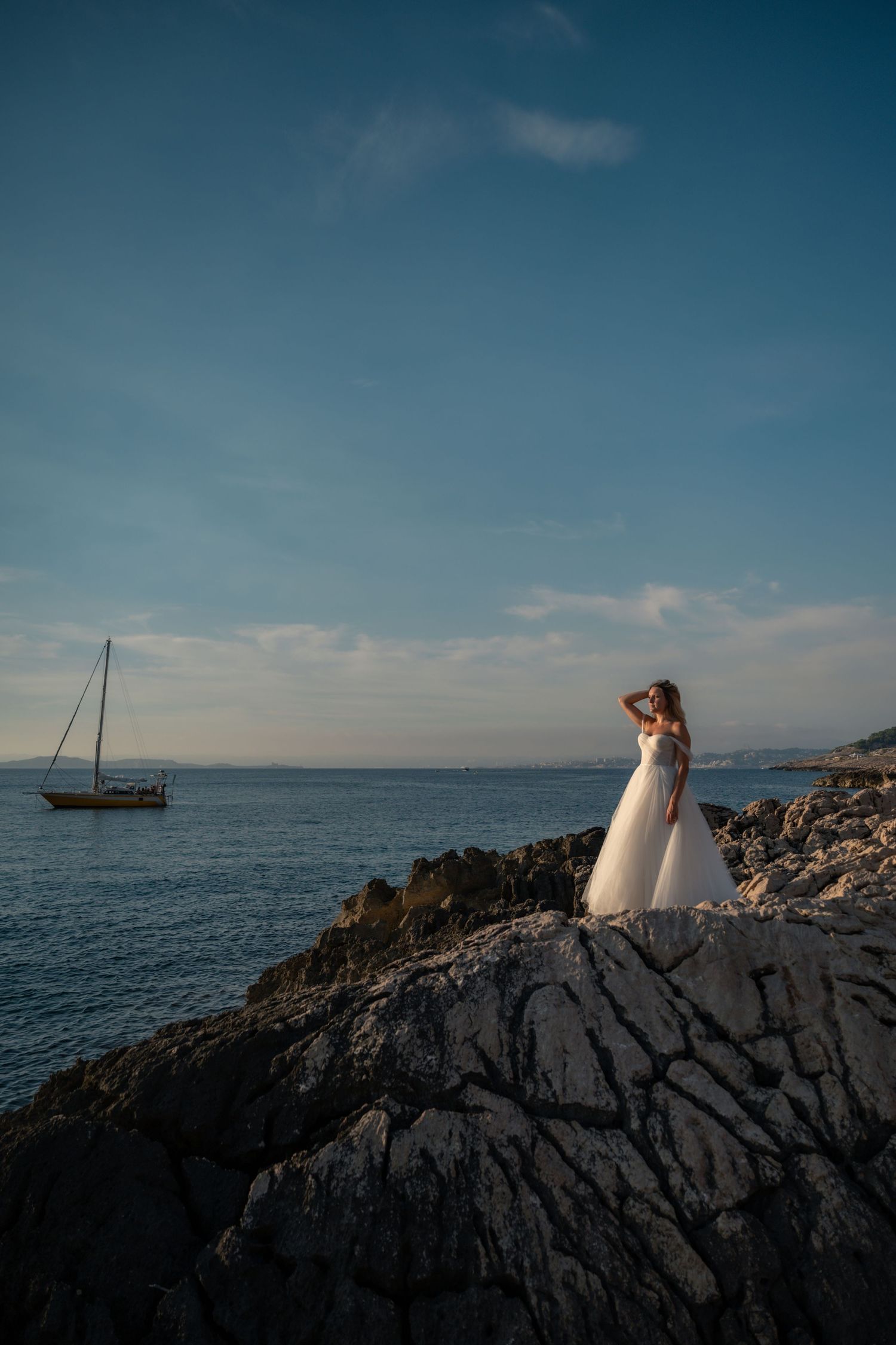 Bride in white gown standing on rocky coastal cliffs with sailboat on blue sea.