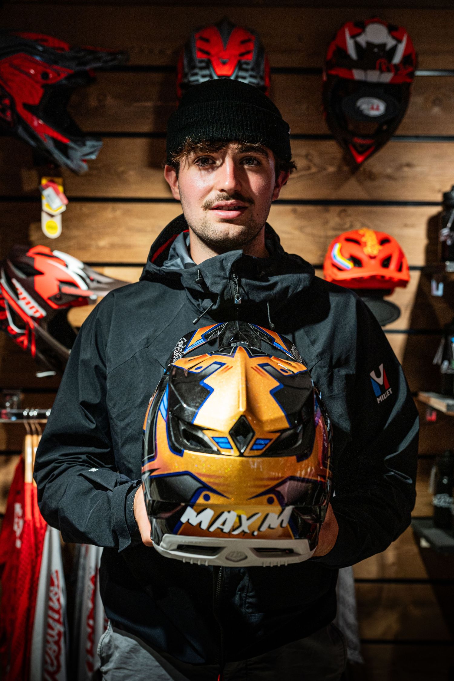 Young man holding a custom painted orange MAXIM motorcycle helmet in a helmet shop.