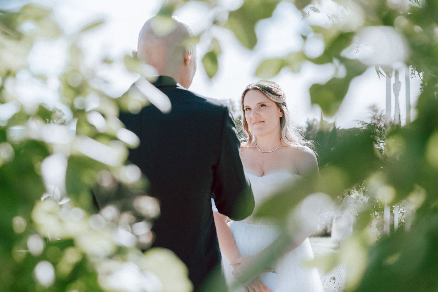Bride in white strapless gown gazes at groom during intimate outdoor wedding ceremony framed by leaves.