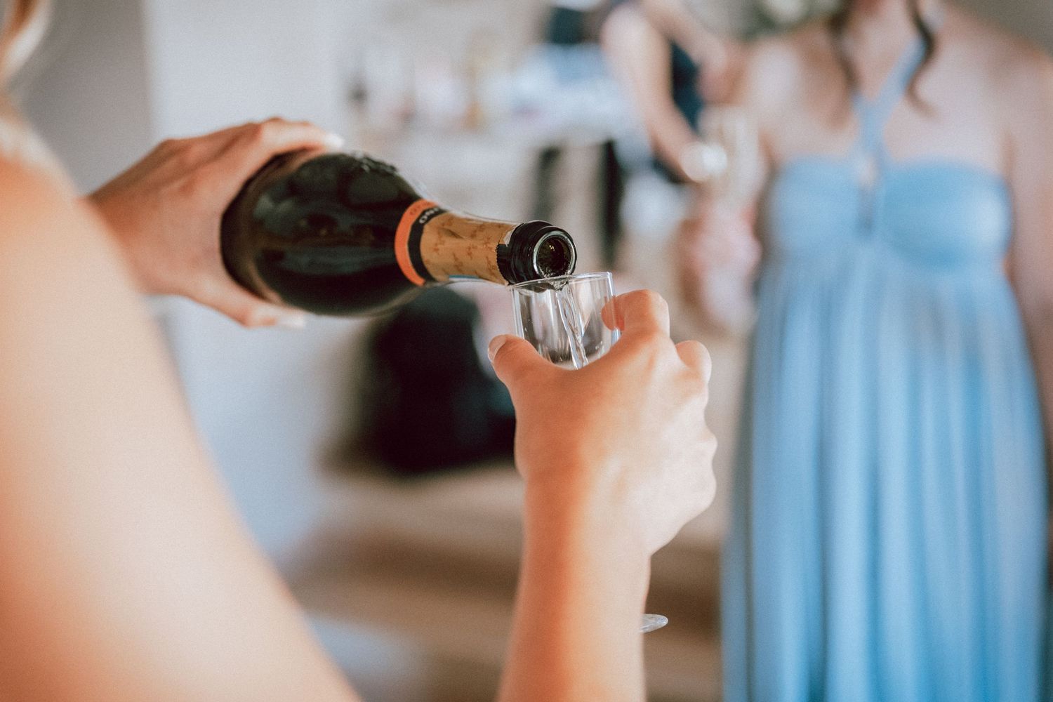 Champagne being poured into a glass at a wedding celebration with bridesmaids in blue dresses in background.