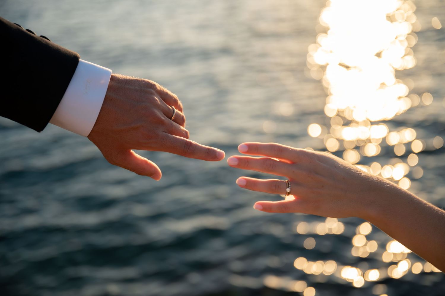 Bride and groom reaching hands toward each other near sparkling ocean water, wedding ring visible on bride's finger.