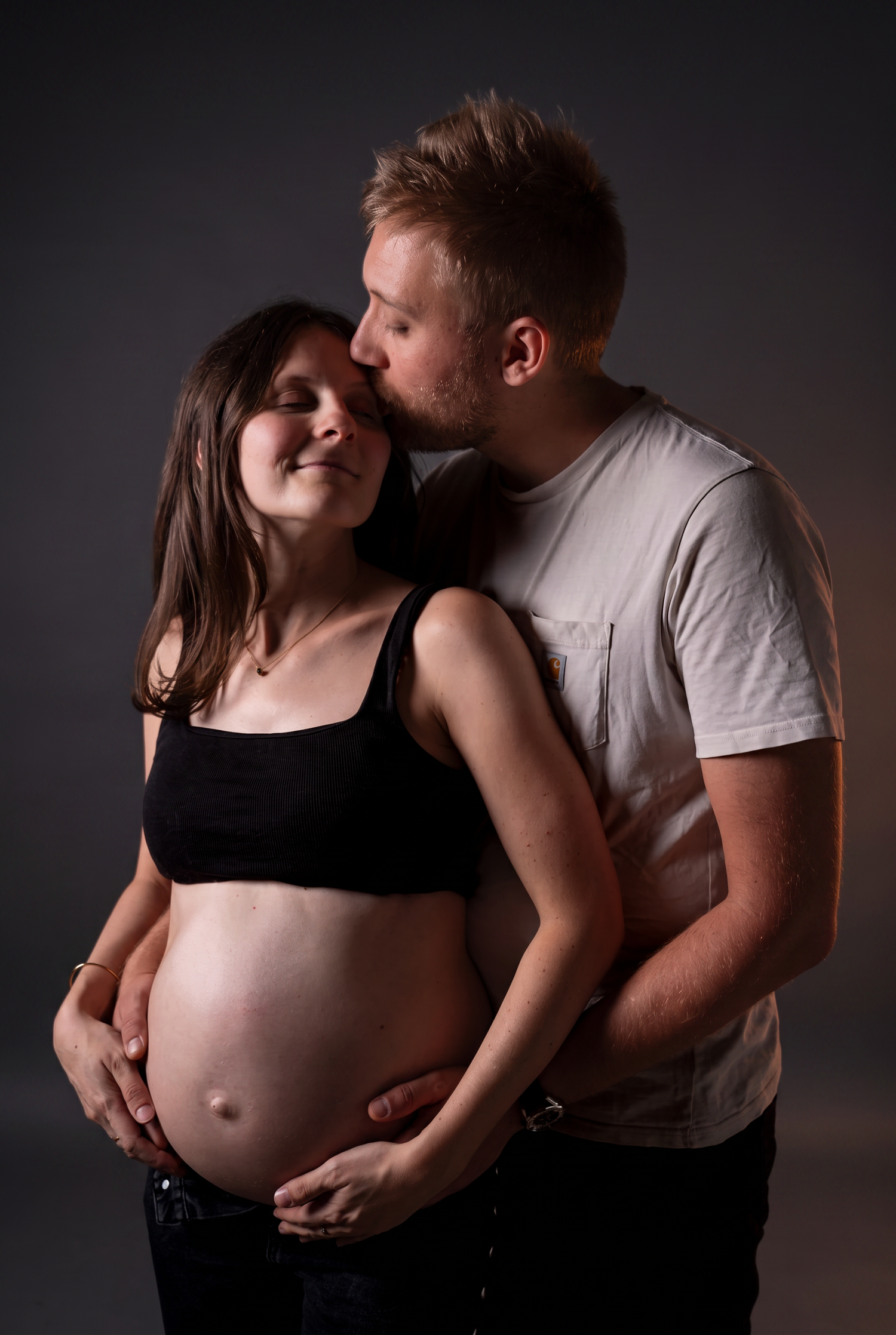 Pregnant couple sharing tender moment, man kissing woman's temple while cradling her baby bump in studio portrait.
