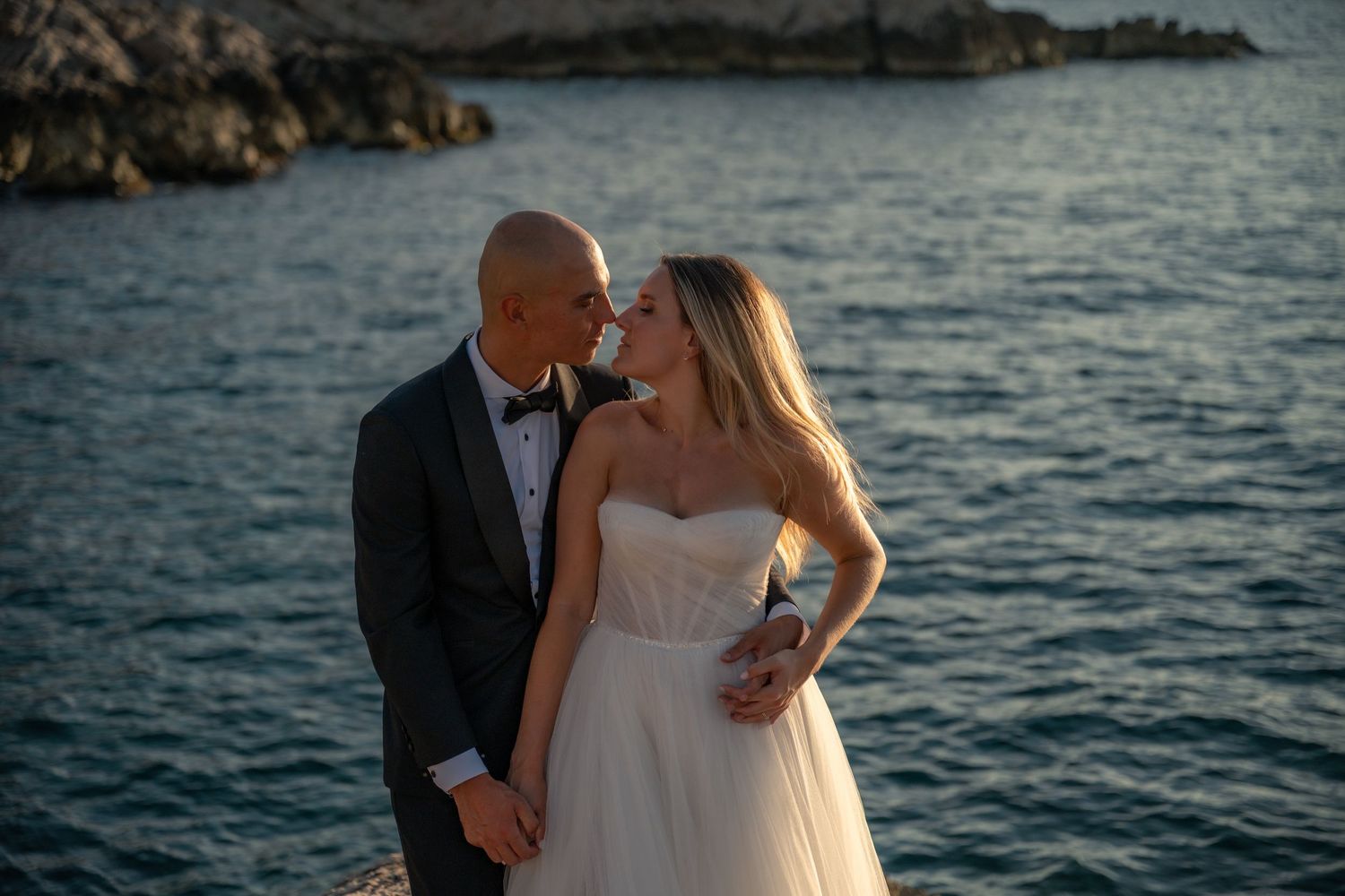 Bride and groom share a romantic moment by the sea, dressed in wedding attire at sunset.