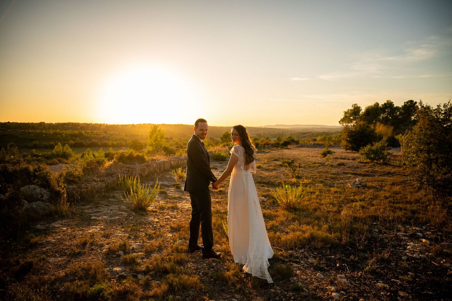Bride and groom holding hands at golden sunset in a rustic outdoor landscape on their wedding day.