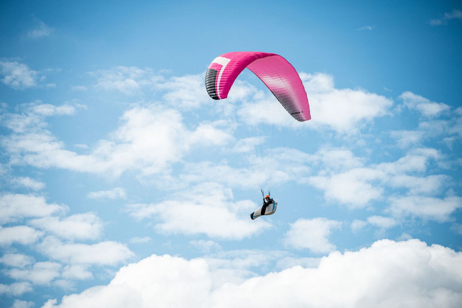 Person paragliding with a pink parachute against a bright blue sky with fluffy white clouds.