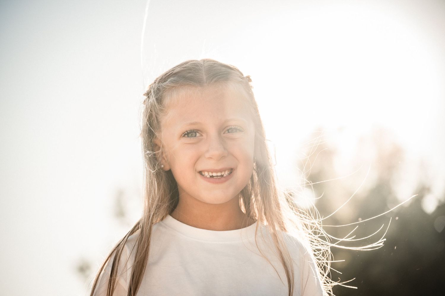 Young girl with long blonde hair smiles brightly in a sun-drenched outdoor portrait.