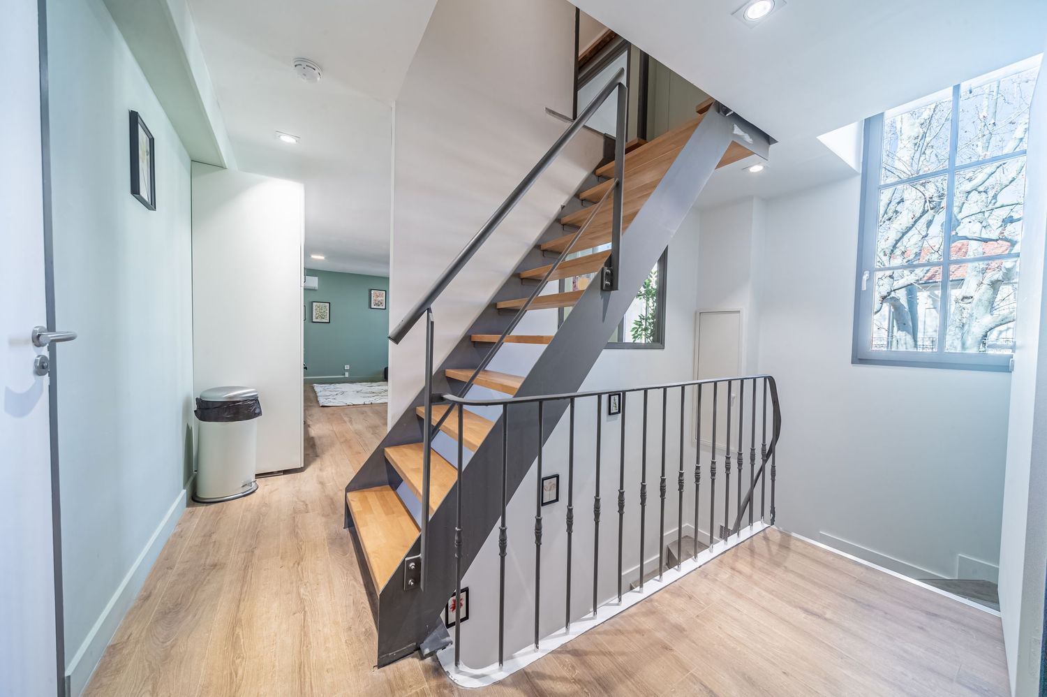 Modern hallway with wooden staircase, black metal railing, light wood floors, and sage green walls in a contemporary home.