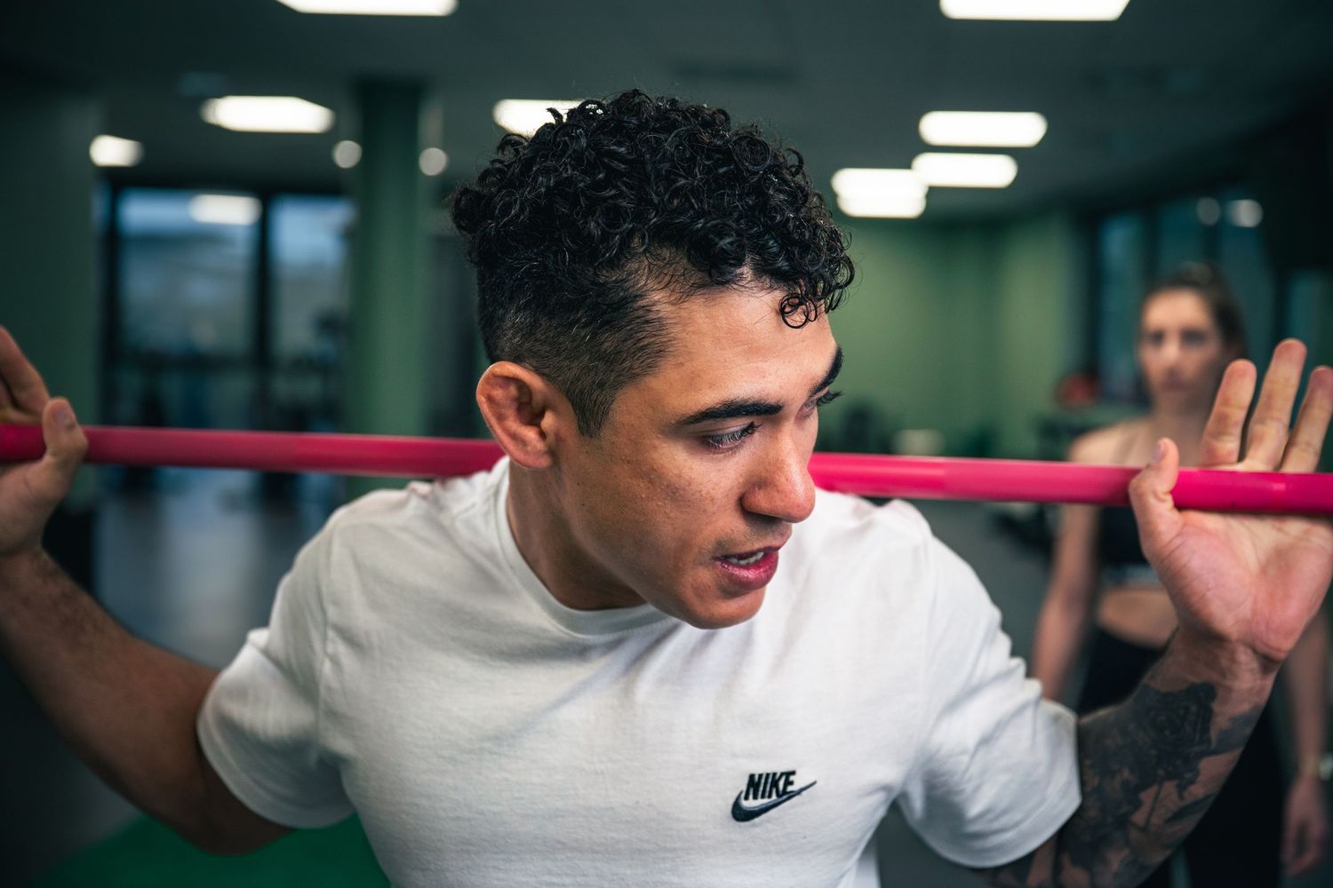 Young man in white Nike shirt exercising with pink resistance bar in gym.