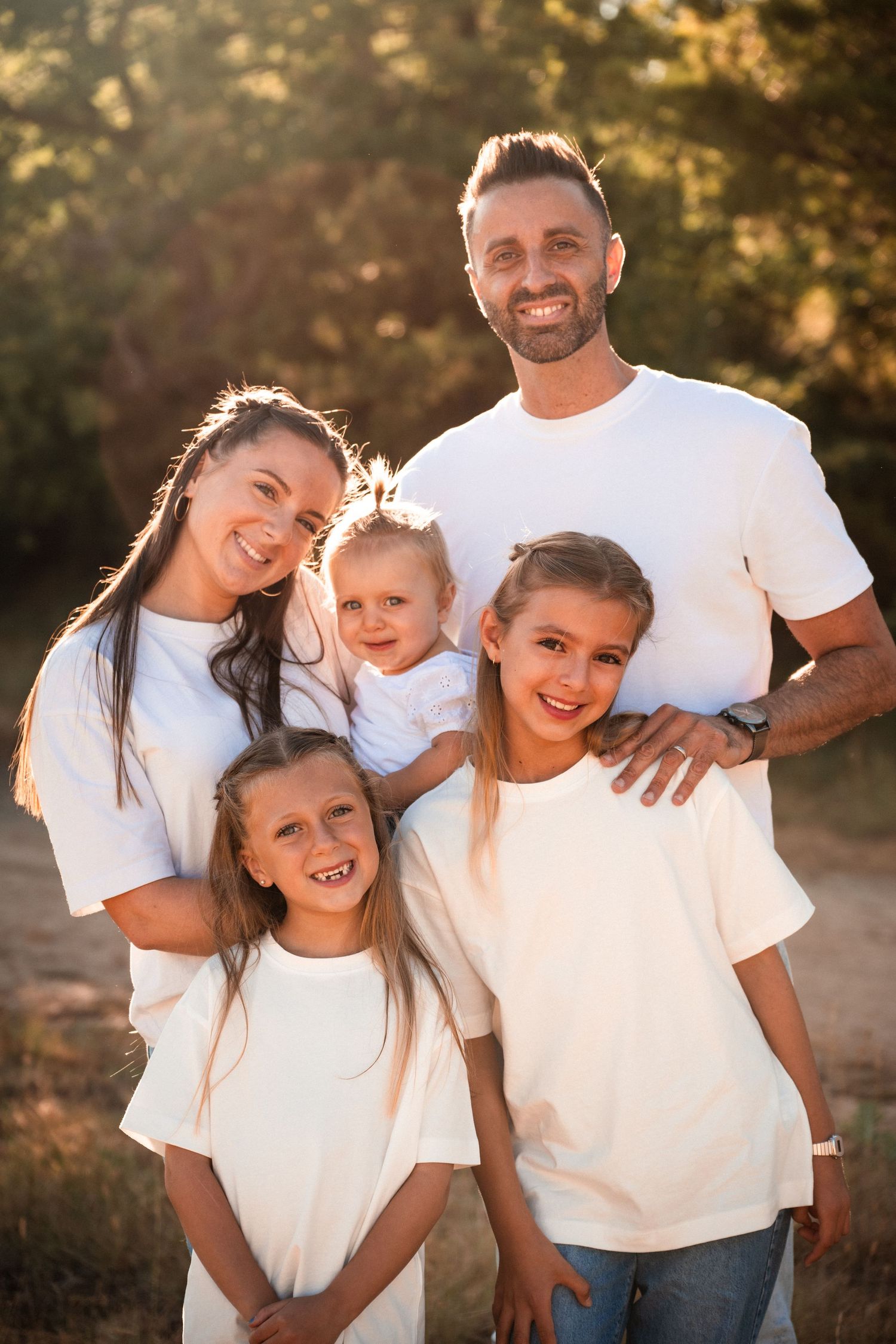 Happy family of five in matching white t-shirts posing outdoors in golden sunlight, parents with three young daughters smiling.