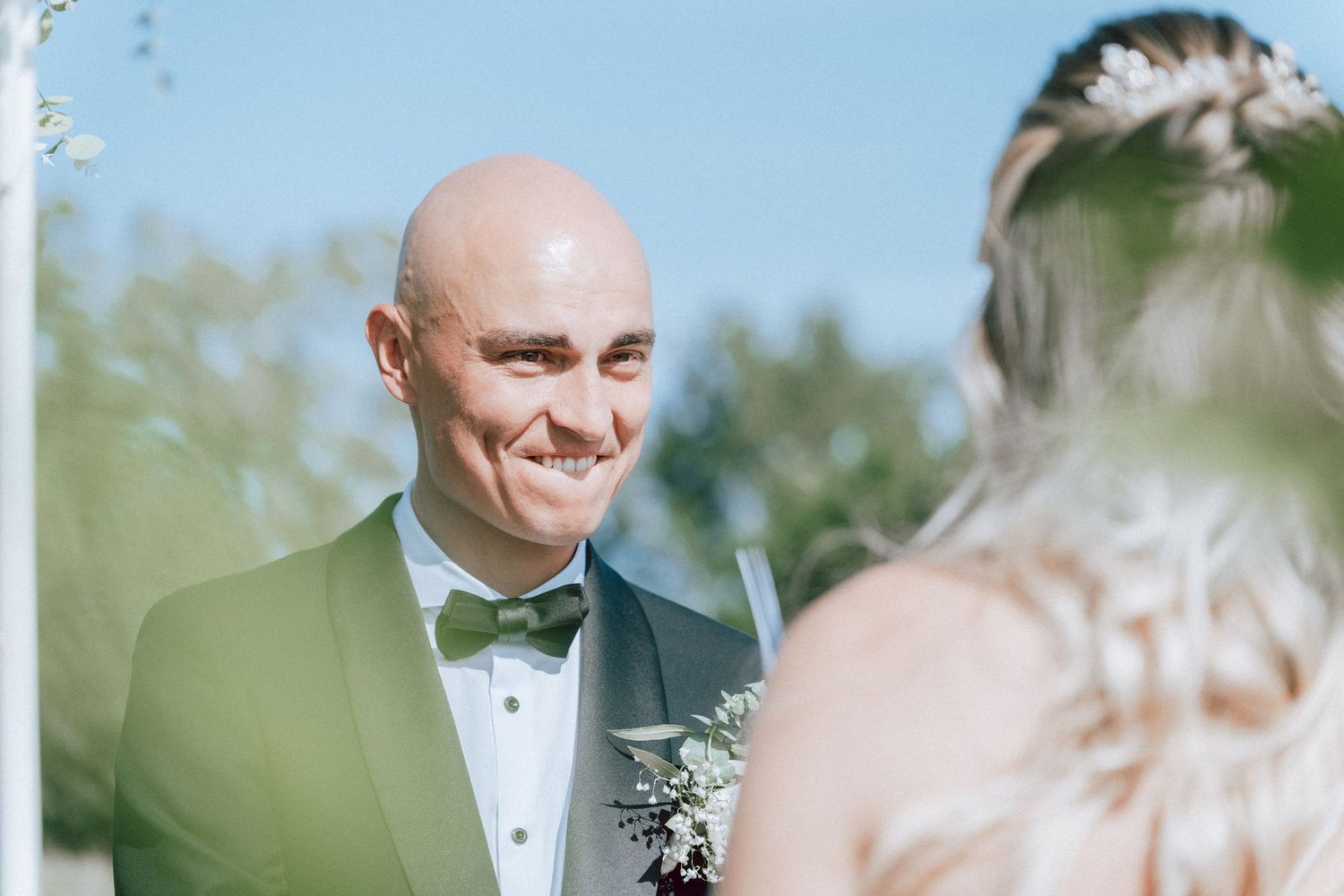 Bald groom in teal tuxedo smiling lovingly at bride during outdoor wedding ceremony.