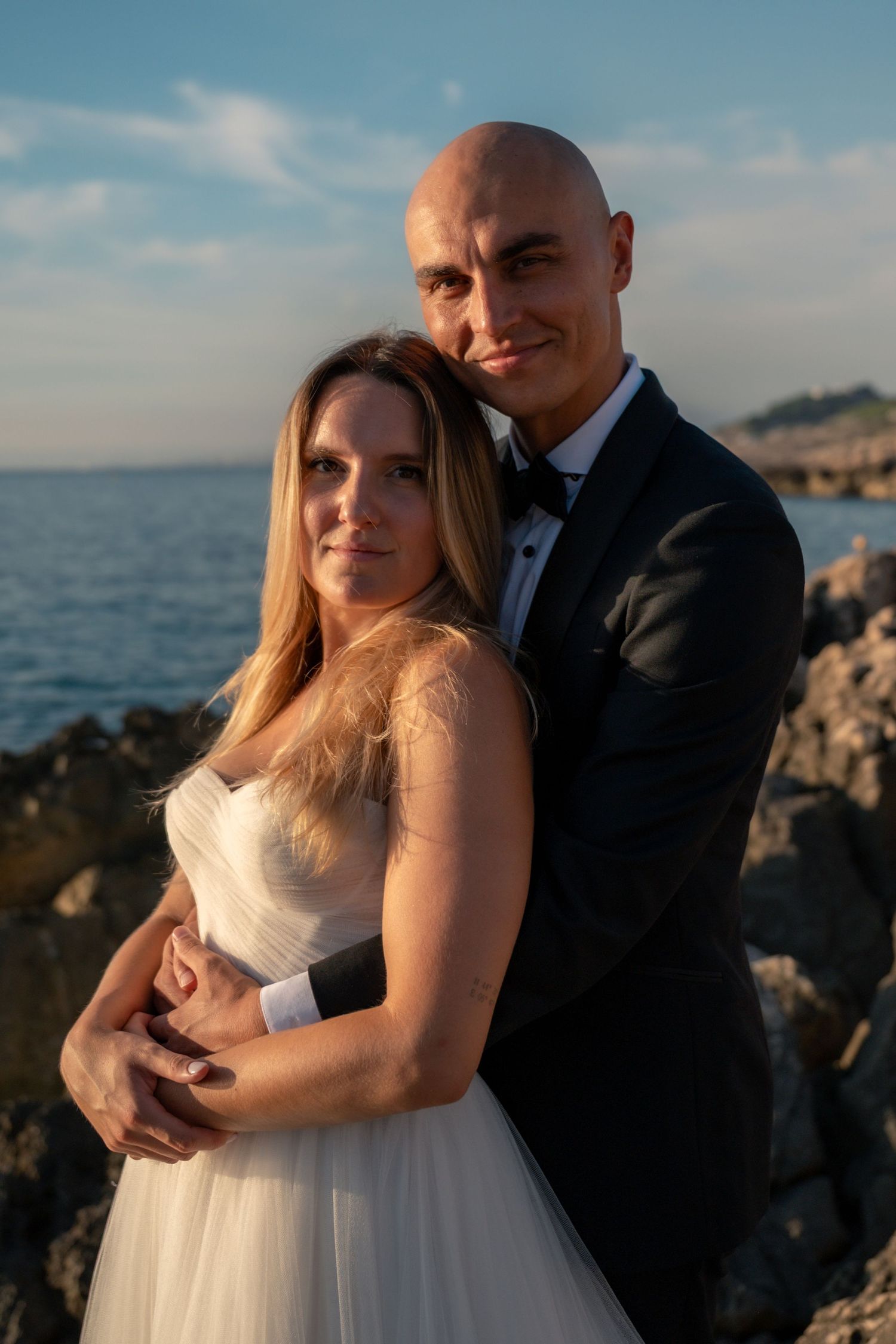 A bride and groom embrace on rocky seaside cliffs at sunset, she in white gown, he in black tuxedo.