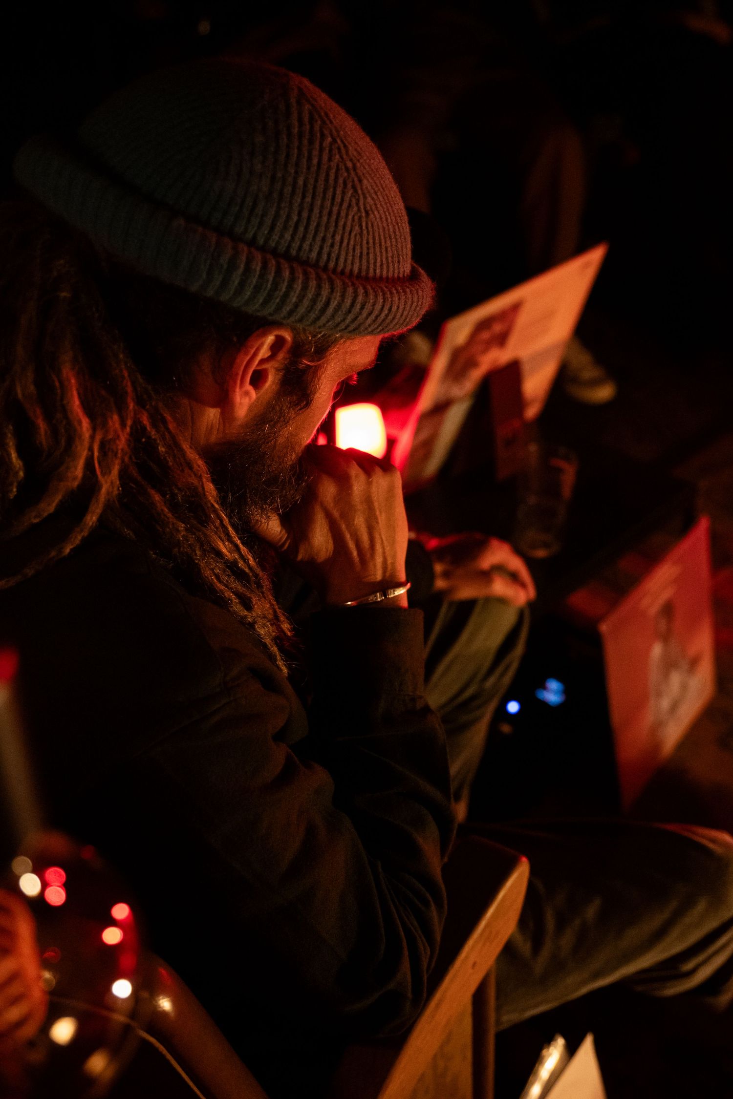 Man with dreadlocks and beanie hat reading by candlelight in a dimly lit venue.