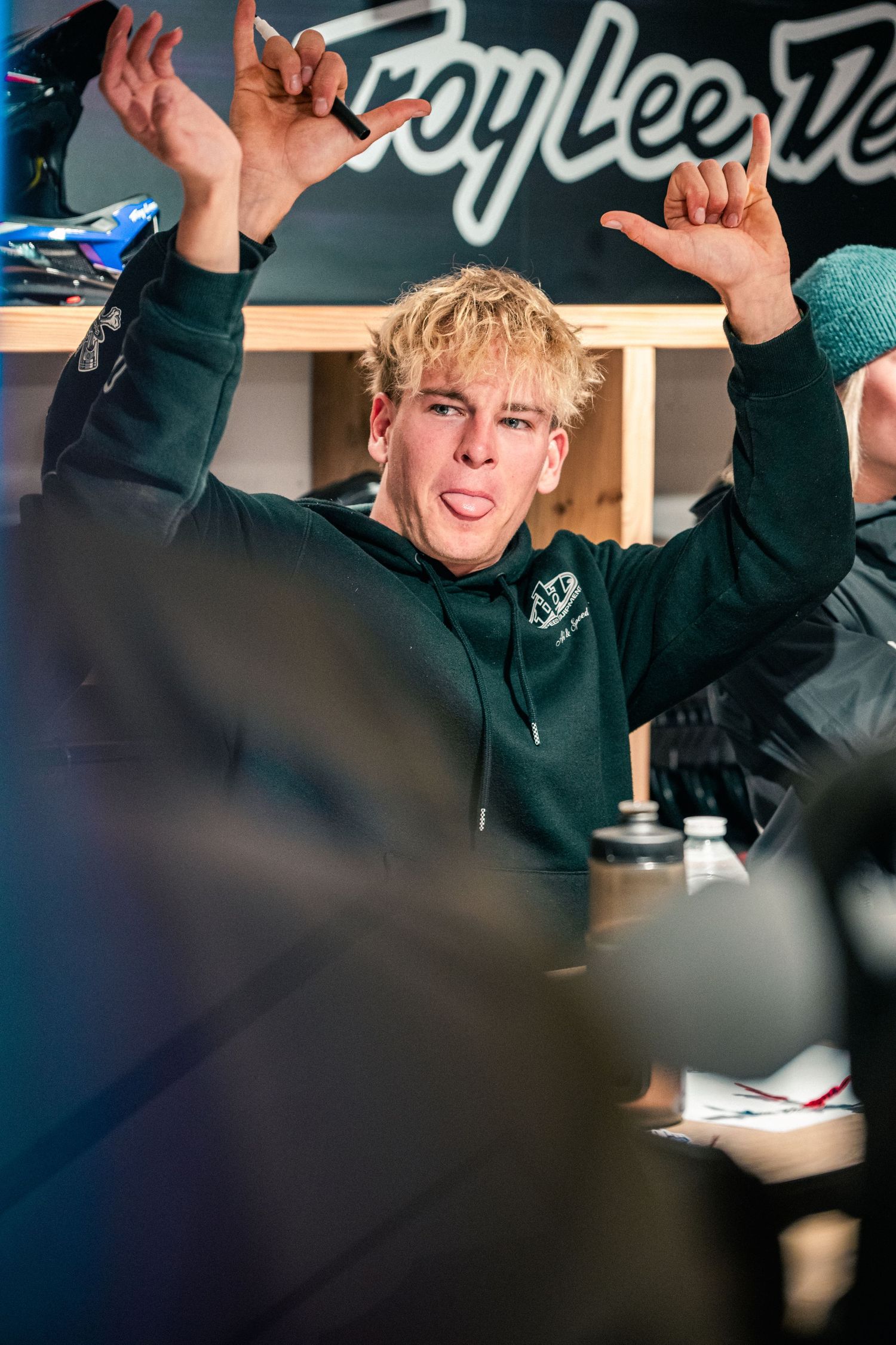 Young man in green hoodie raises hands triumphantly at Troy Lee Designs event, making rock gesture with tongue out.